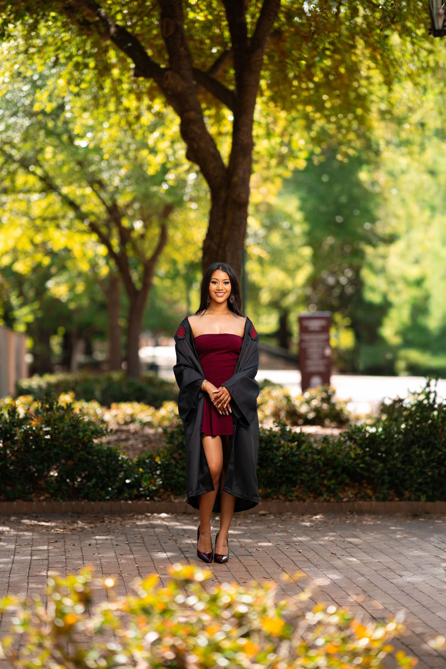 Smiling graduate in burgundy dress and gray gown walks along tree-lined brick path outdoors.