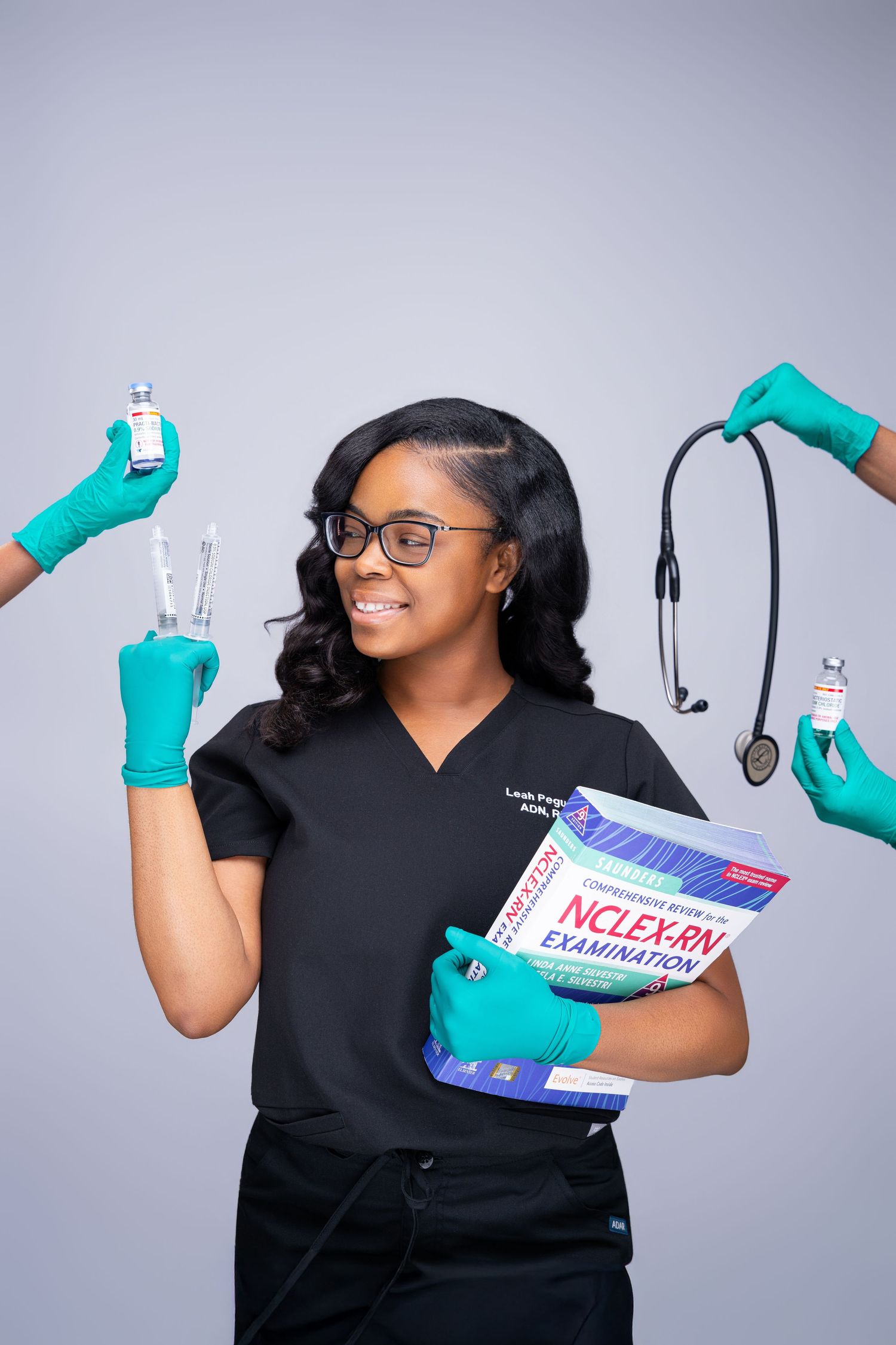 Smiling nurse in black scrubs holds NCLEX-RN study book surrounded by hands with medical tools and stethoscope.