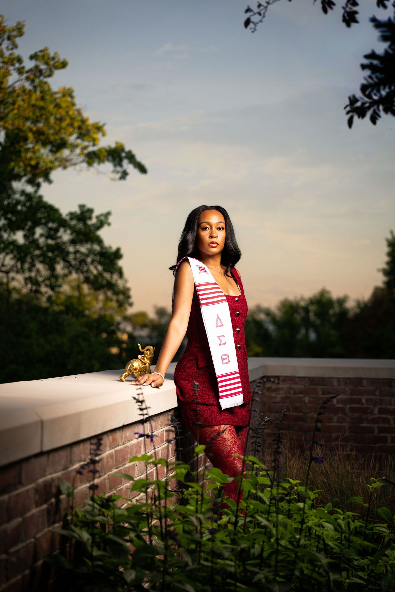 A young woman in a red dress wearing a Delta Sigma Theta graduation sash poses outdoors at sunset.