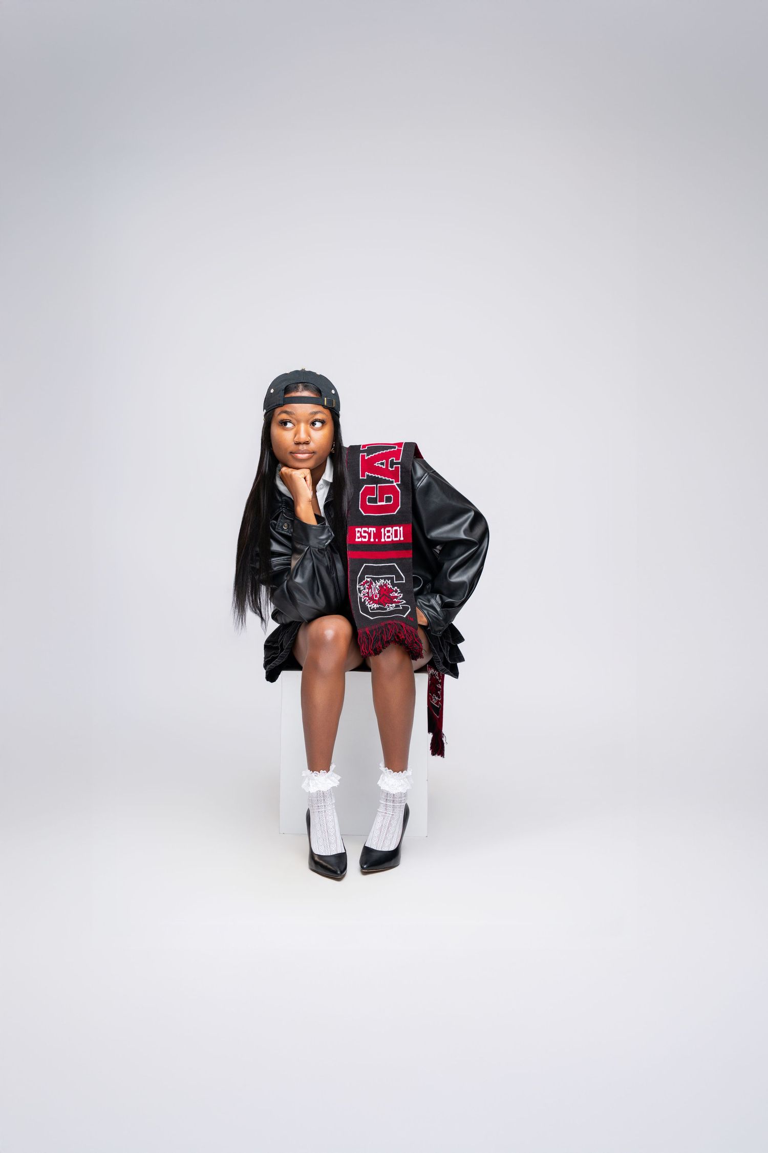 Young woman in black leather jacket, red collegiate scarf, white socks, and cap sits posing on white background.
