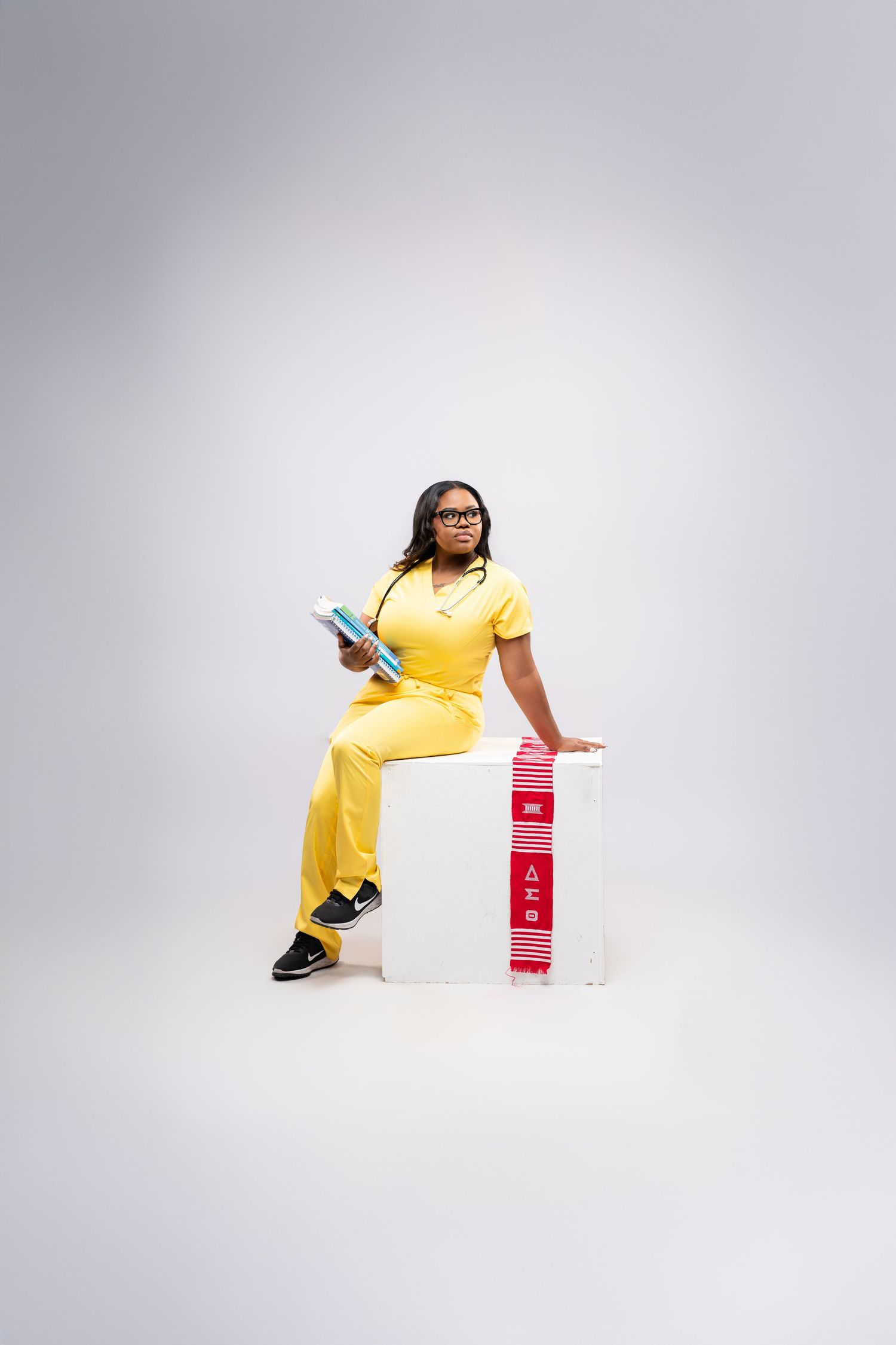 Woman in yellow scrubs sitting on white cube with red Delta Sigma Theta paraphernalia, holding books.