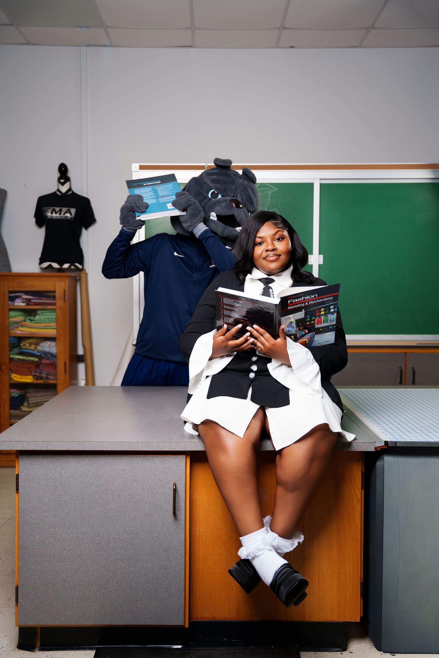 A smiling student in uniform sits on a desk holding a book beside a school mascot in a classroom.