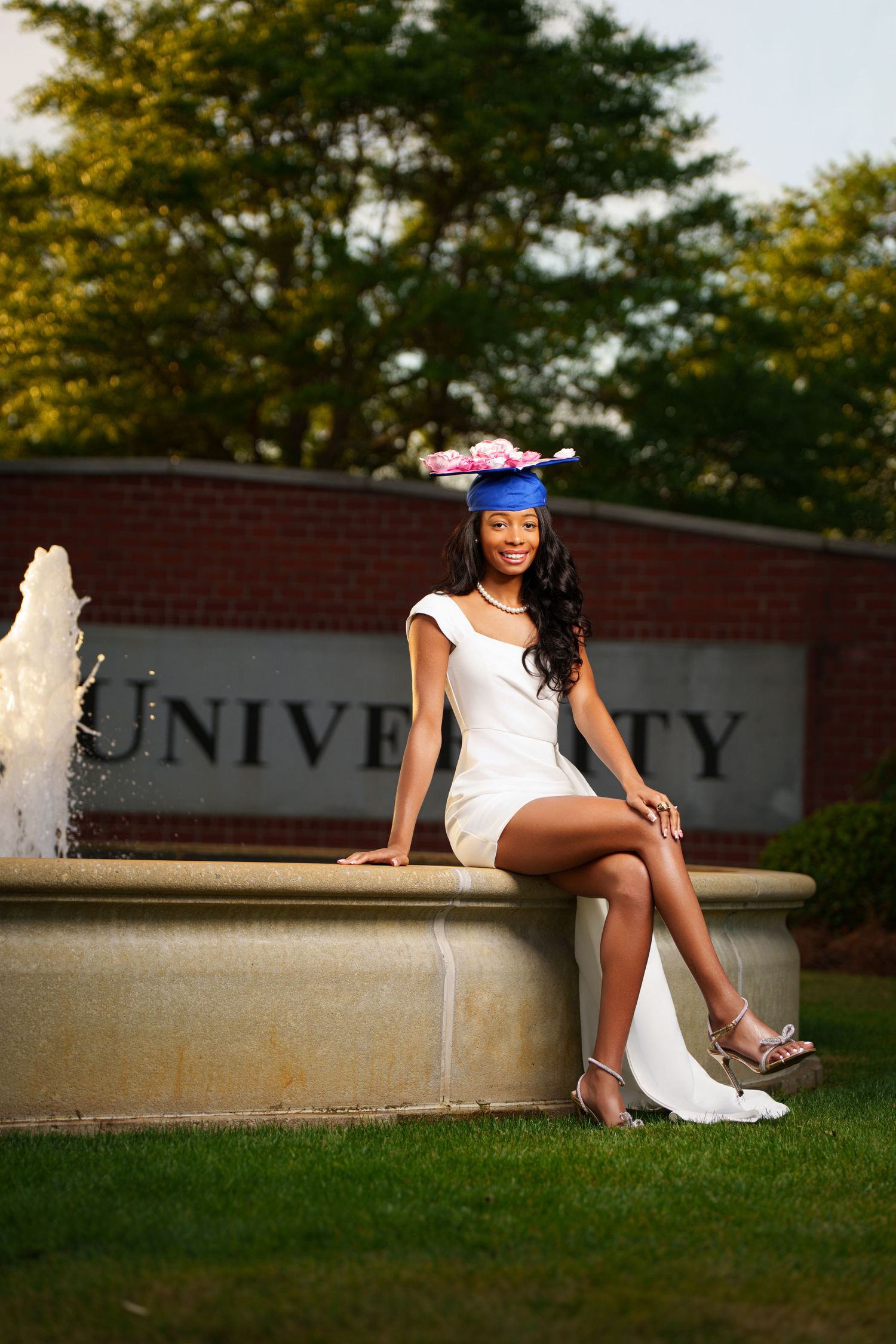 Young woman in white dress and blue graduation cap sitting on university fountain ledge, smiling outdoors.