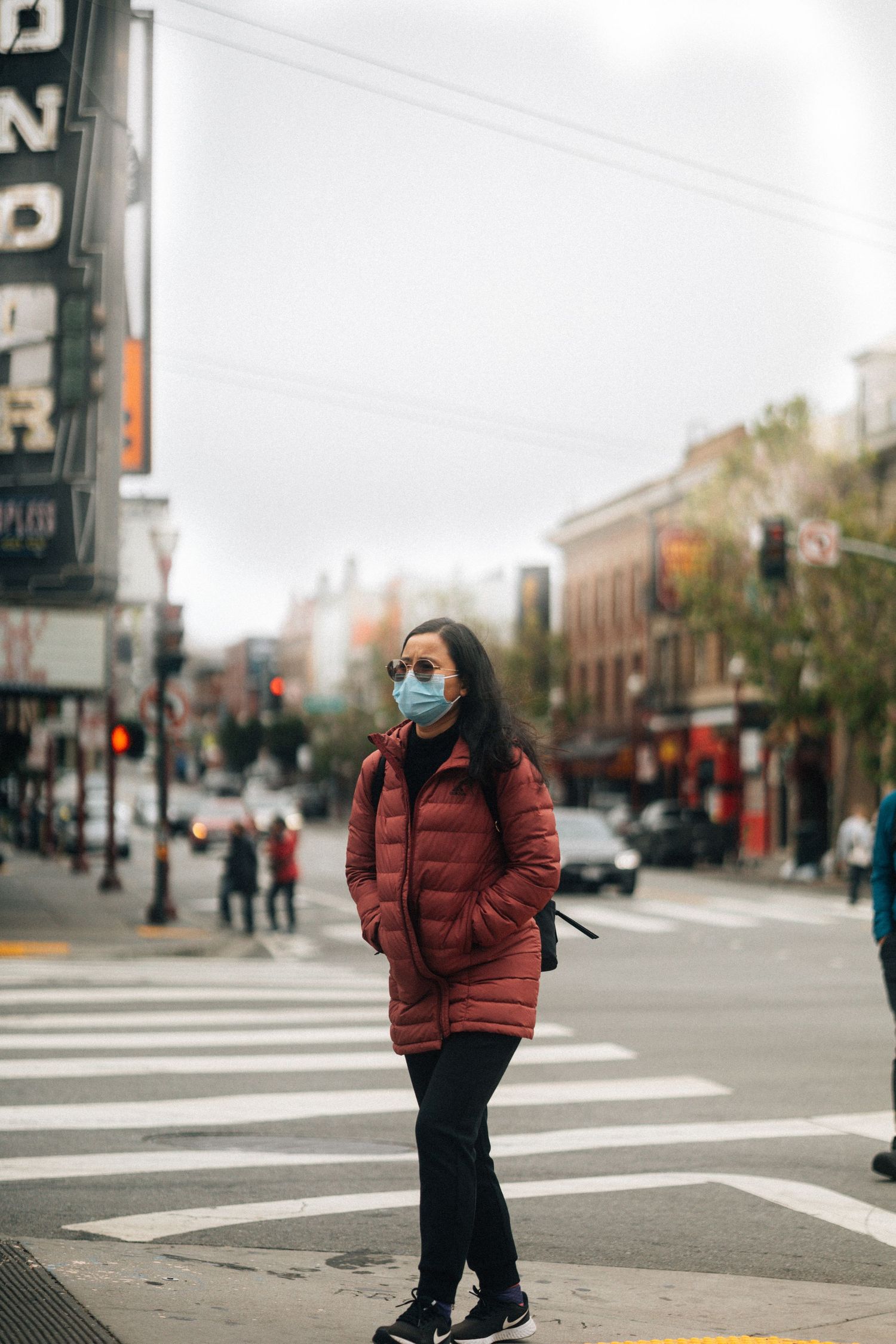 Person wearing red puffy winter coat and face mask stands at city intersection on cloudy day.