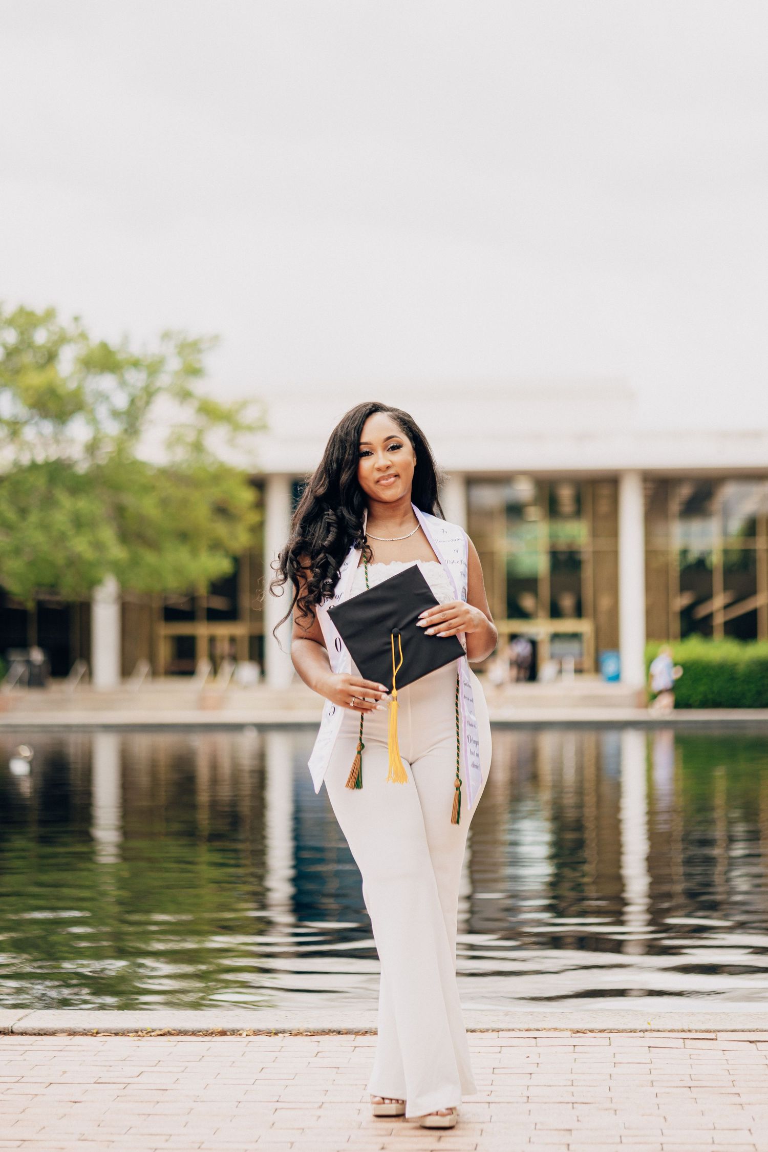 Graduate in white dress poses by reflective pool.