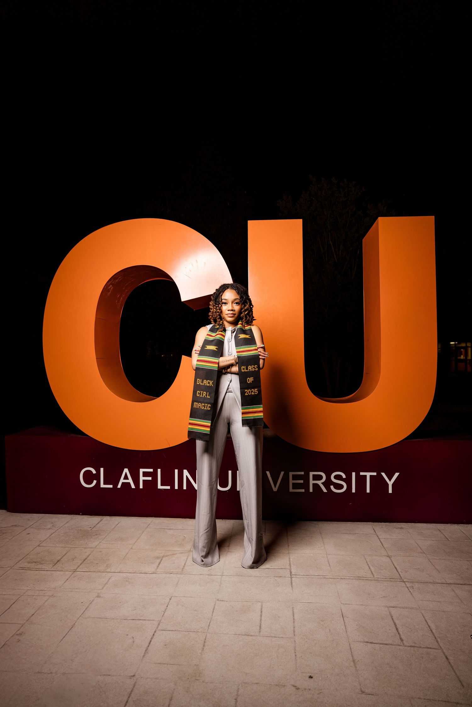 Graduate stands confidently in front of large orange Claflin University sign.