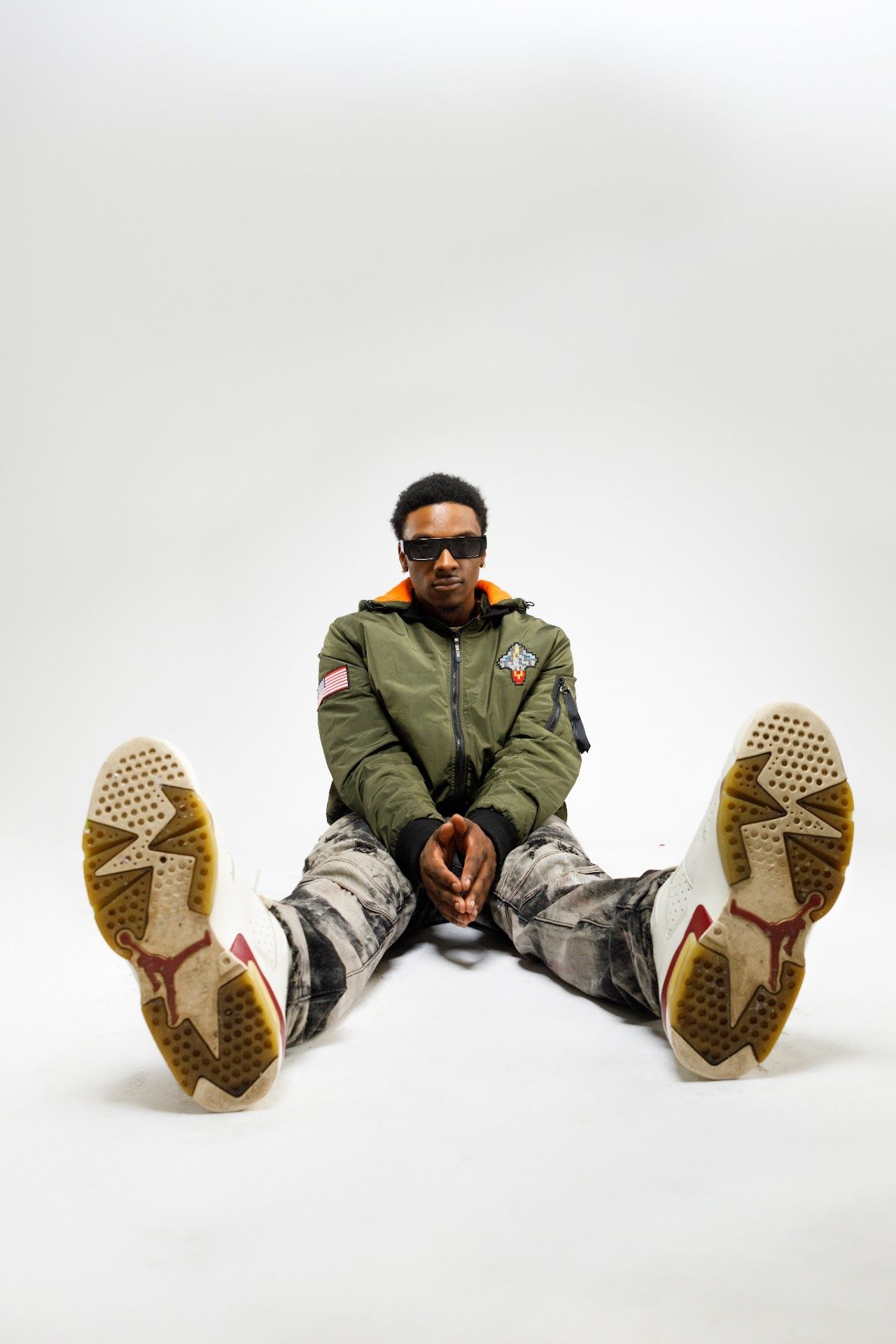 Person in olive bomber jacket and camo pants sitting with legs spread on white background showing shoe soles.