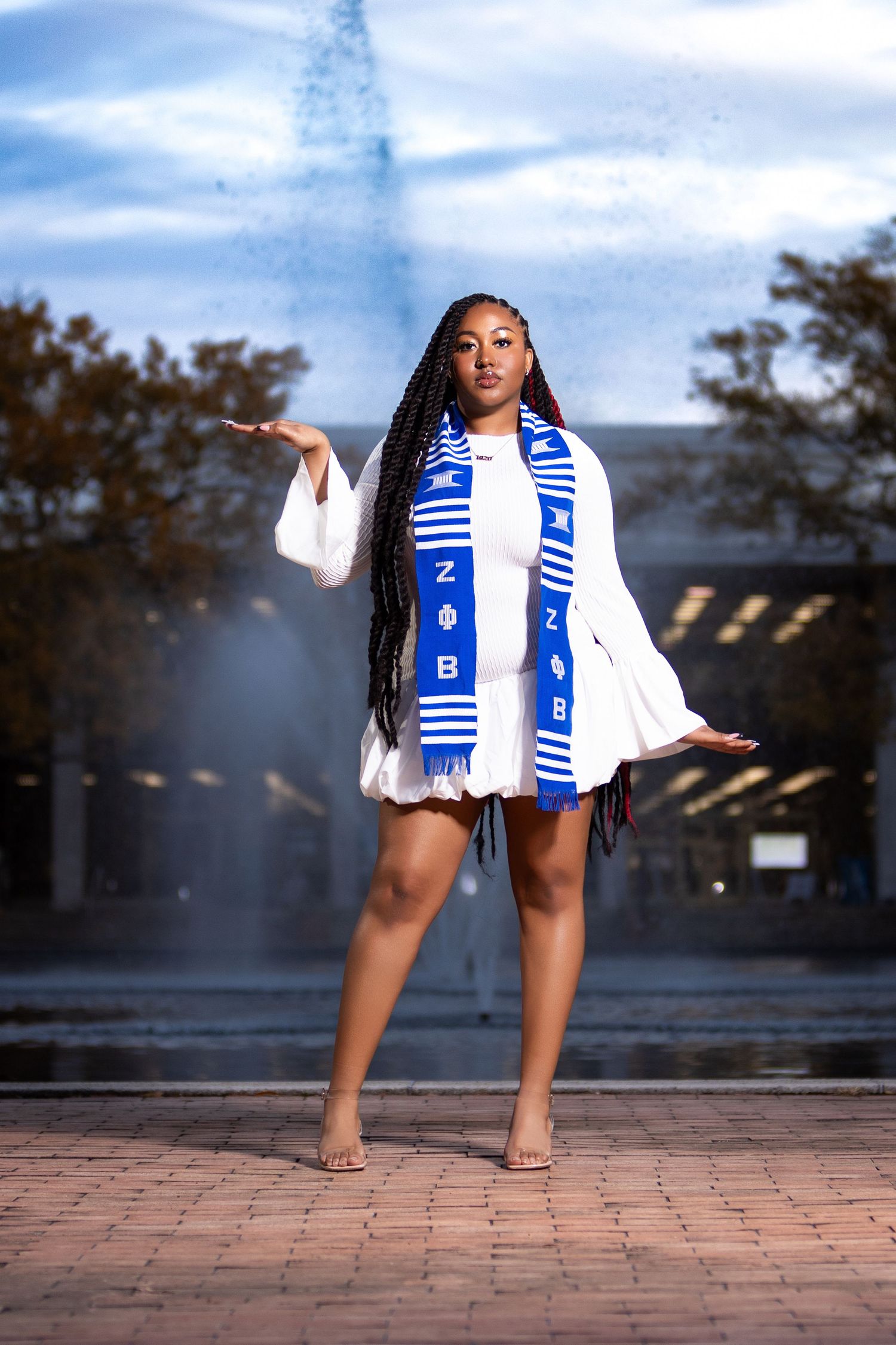 Woman in white dress with blue and white striped scarf poses outdoors on brick pavement.