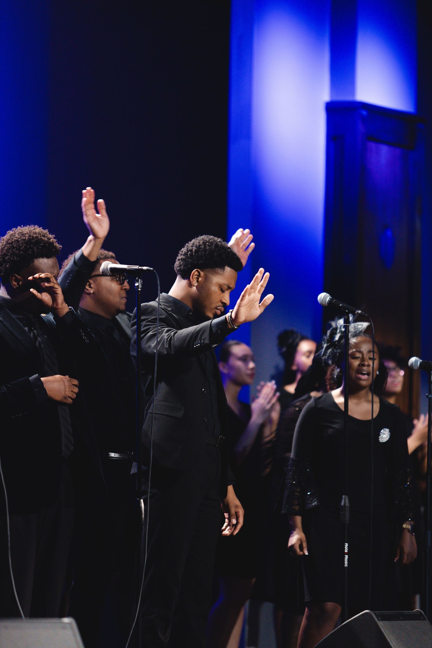 Gospel choir with raised arms performing against blue-lit backdrop.
