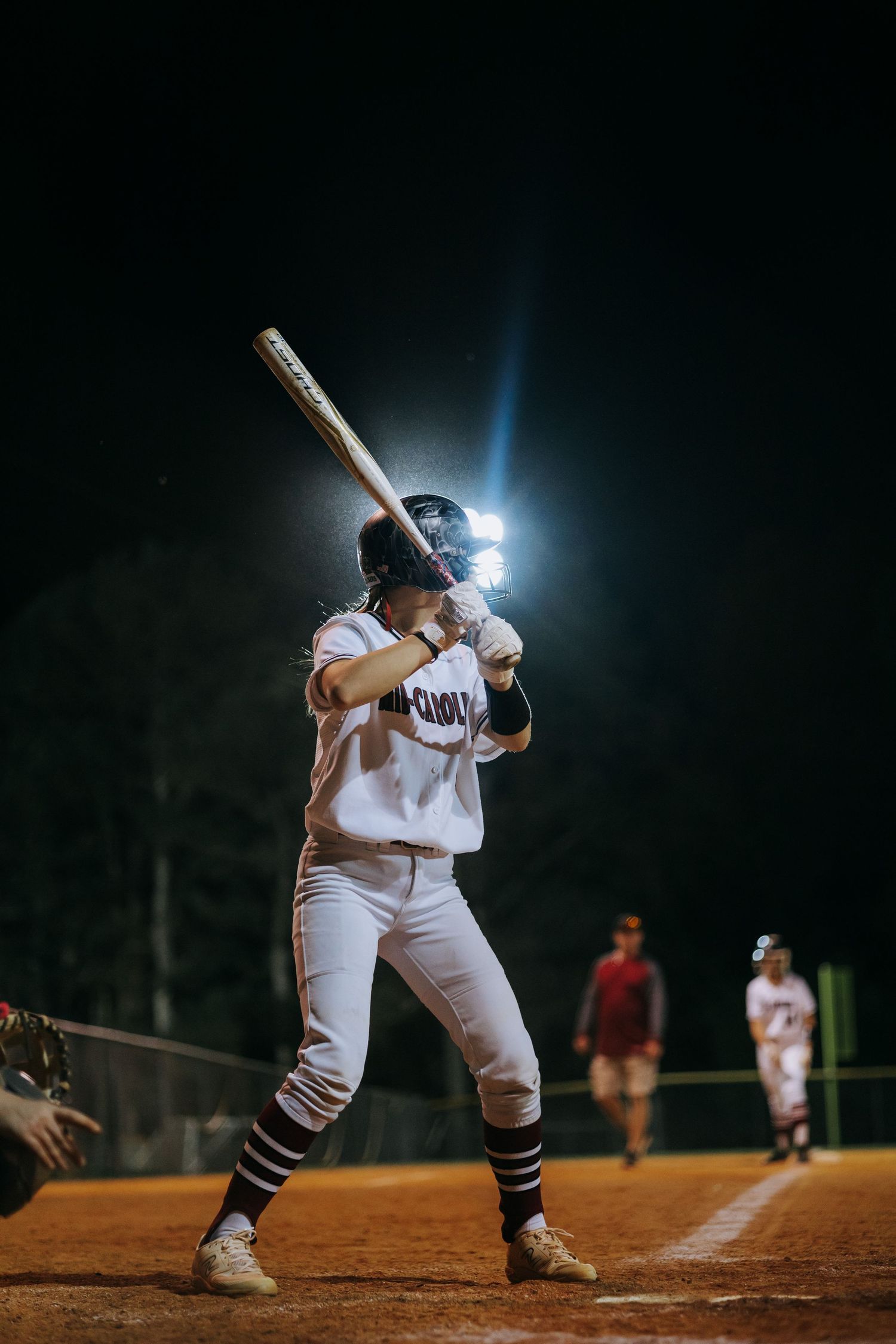 Baseball player at bat under dramatic stadium lighting on dirt field at night.