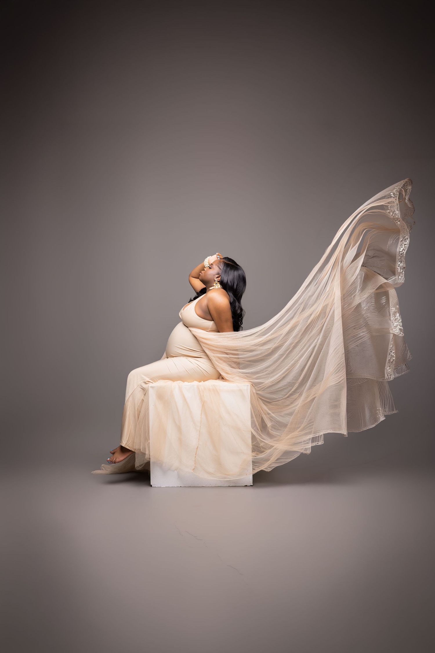 Pregnant woman in flowing beige fabric gown seated on white cube, fabric billowing gracefully in studio setting.