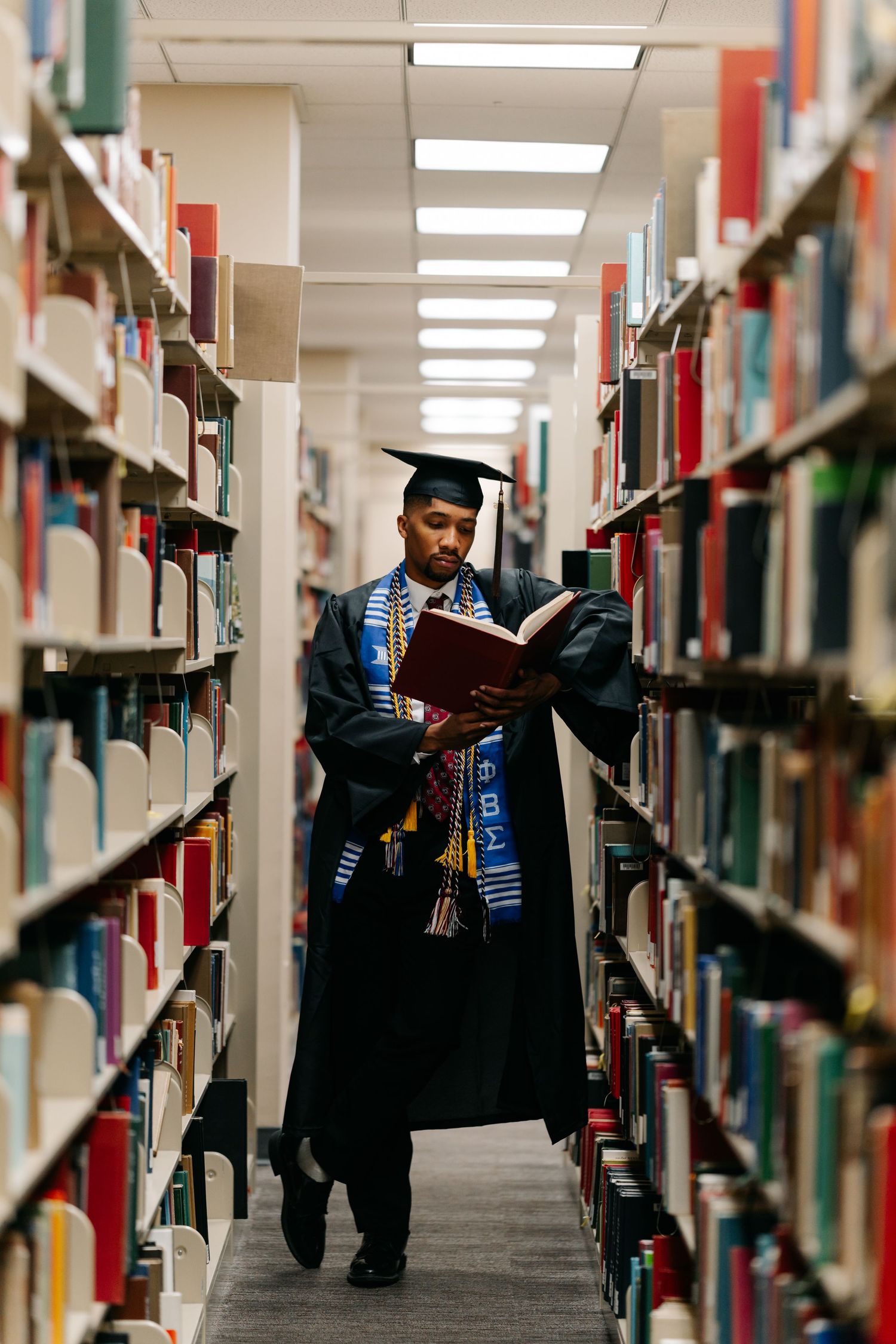 Graduate in cap and gown reads book between library shelves, wearing colorful honor stoles.