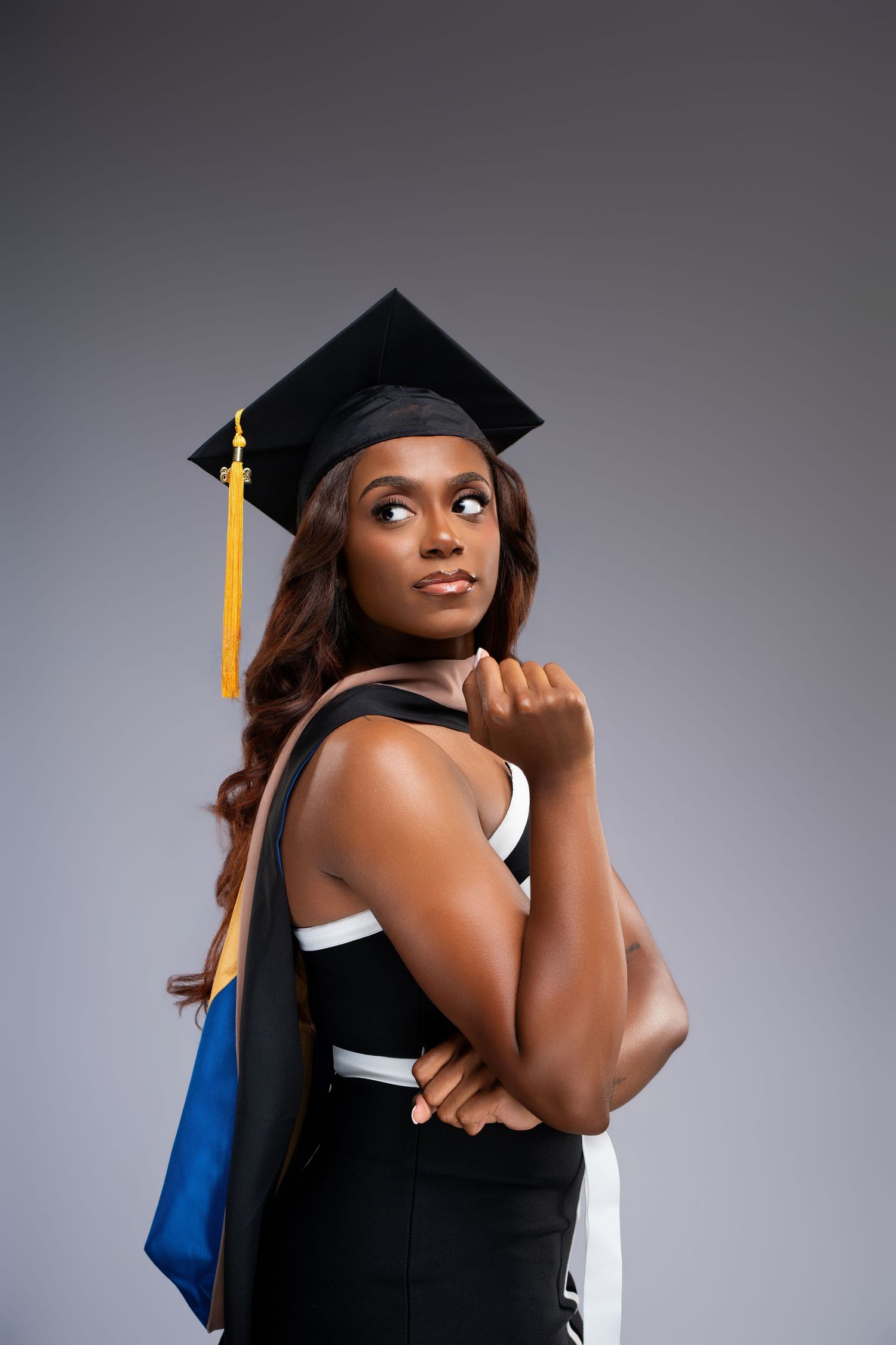 Young Black woman in graduation cap and gown posing confidently against gray background.