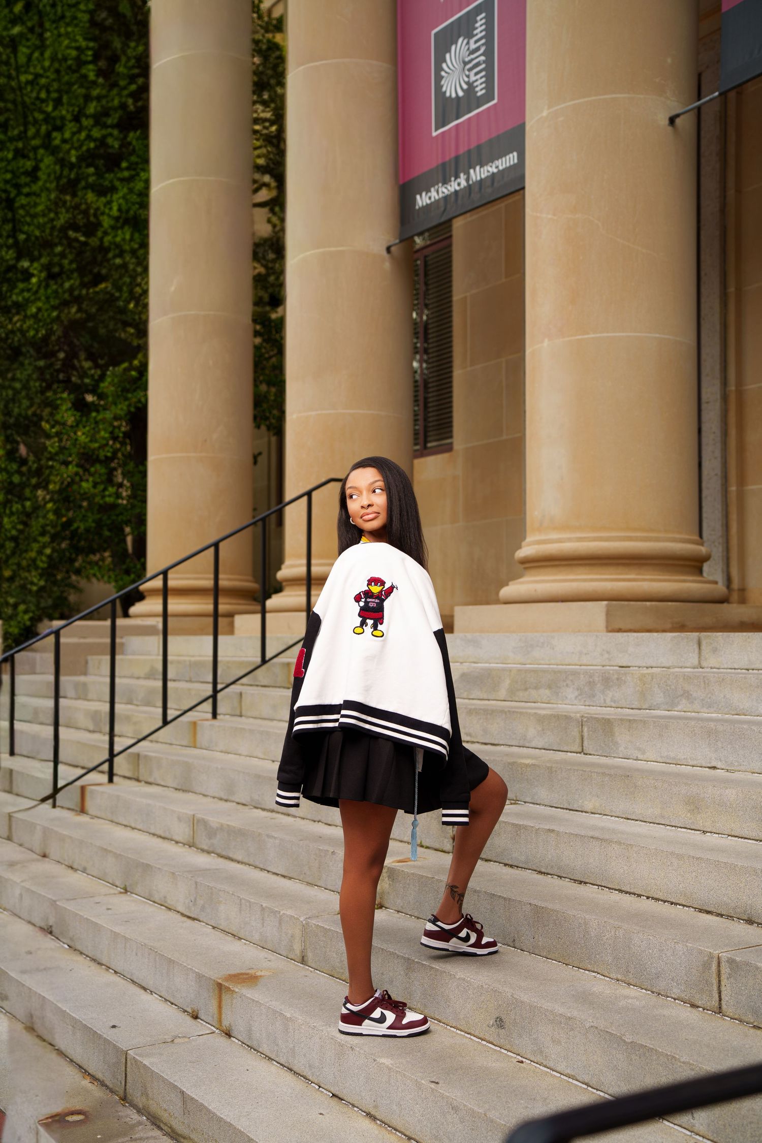 Young woman in white varsity jacket and black pleated skirt poses on stone steps outside McKinrick Museum.