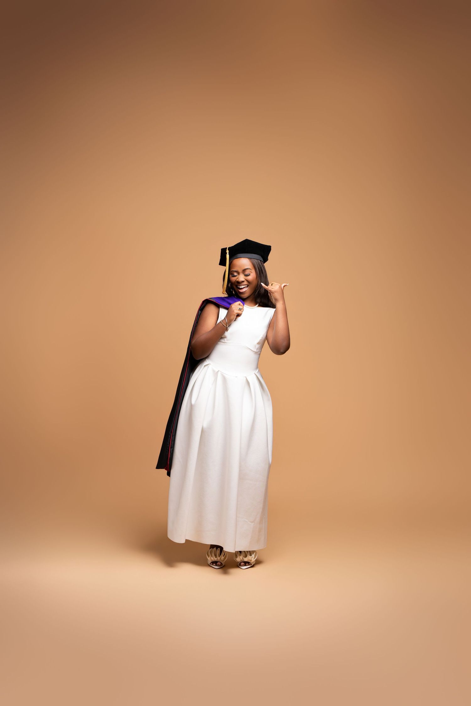 Joyful Black woman in white graduation gown and mortarboard cap celebrating her achievement on beige background.