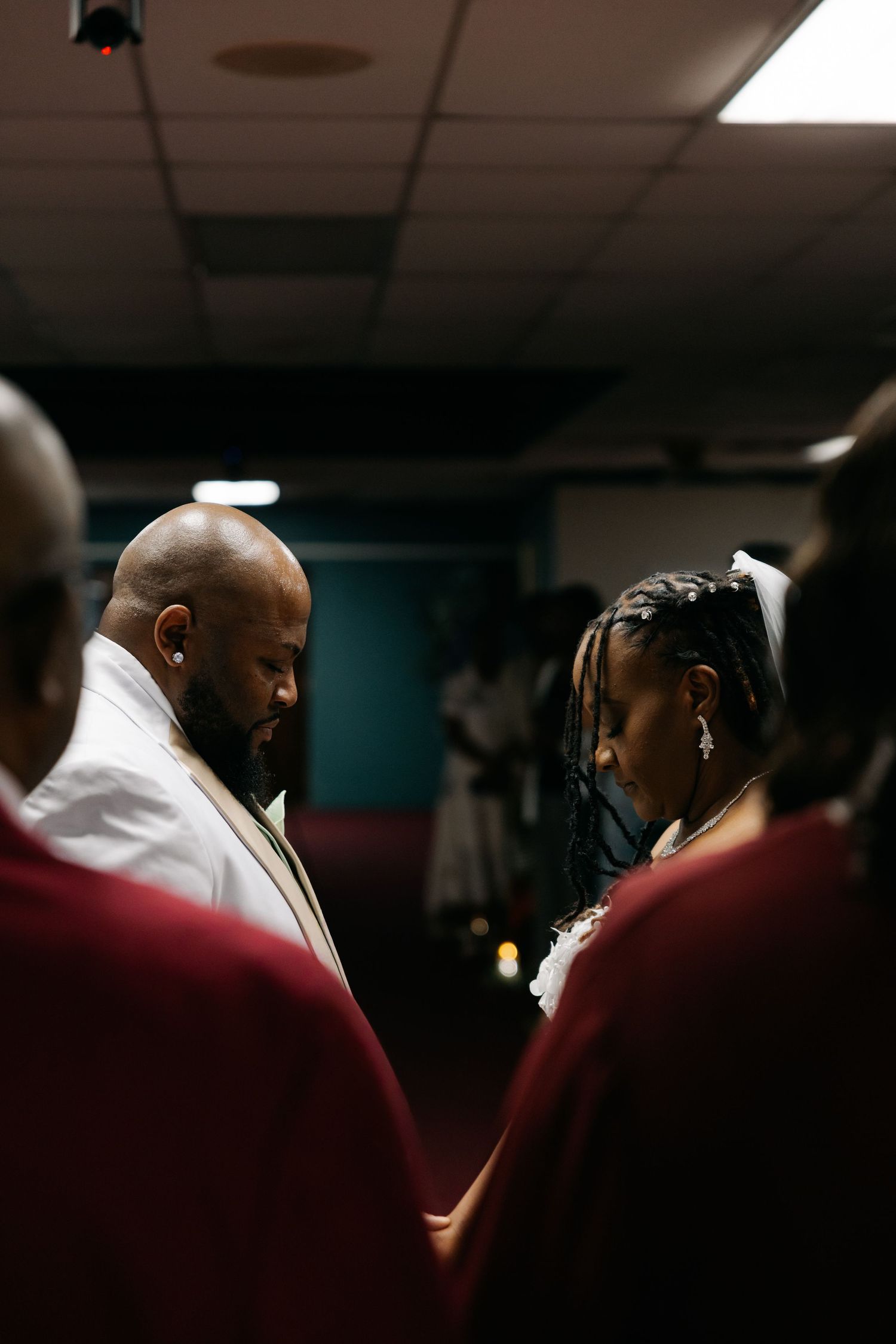 Bride and groom bow heads in prayer during intimate wedding ceremony indoors, surrounded by guests in formal attire.