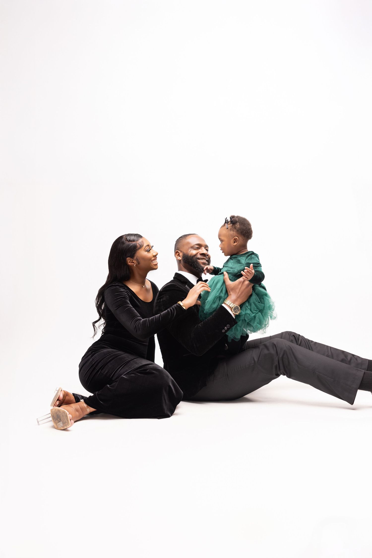 Family portrait with couple in black outfits playing with child in teal outfit on white background.