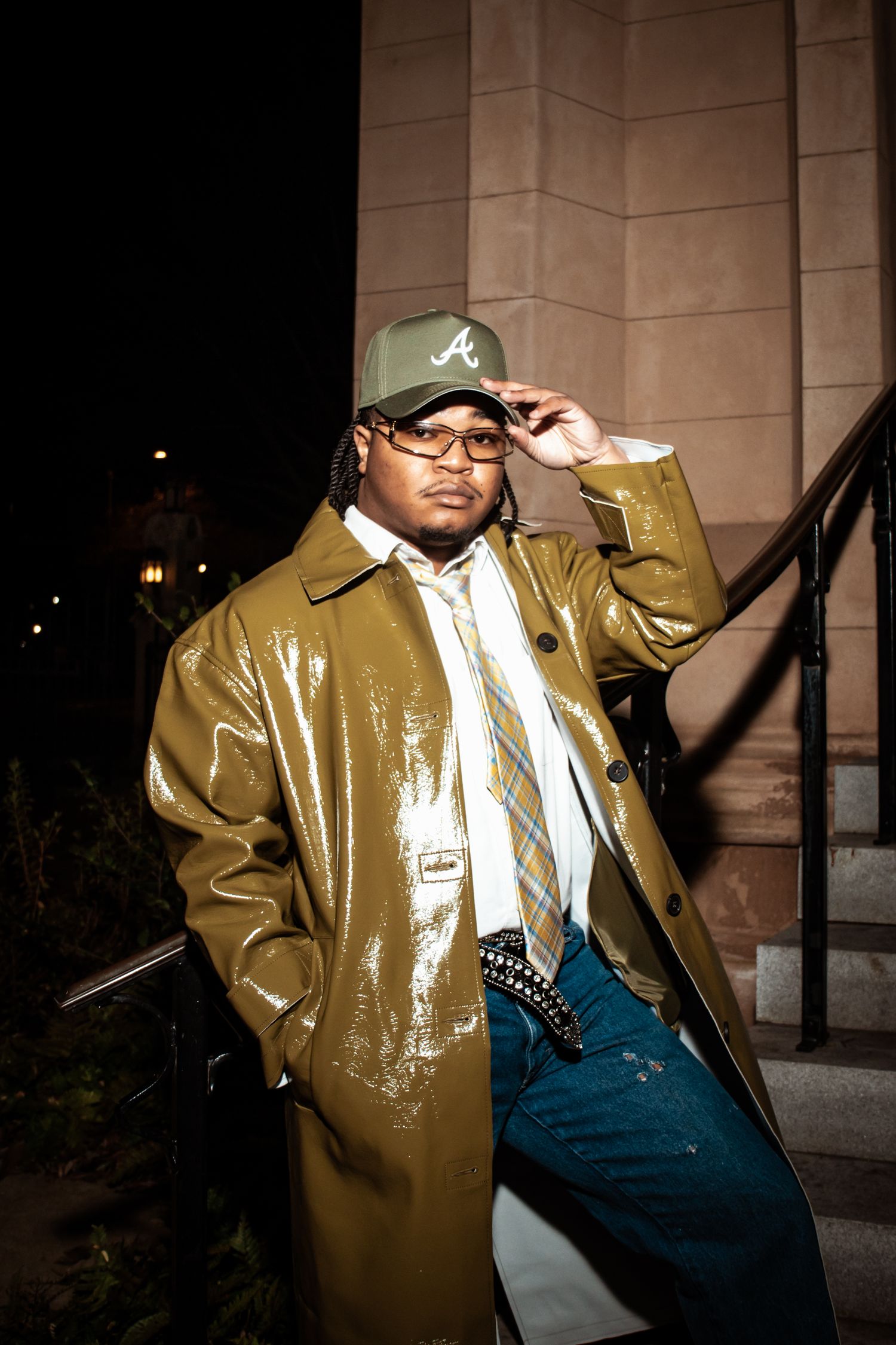 Person wearing a brown vinyl trench coat and Atlanta Braves baseball cap poses against stone building at night.