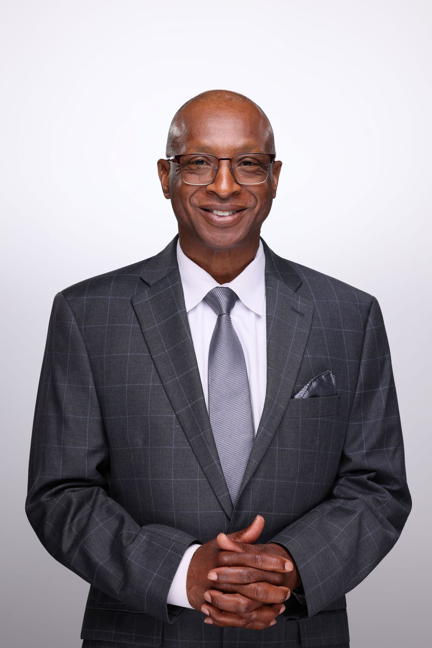 Smiling senior Black businessman in gray plaid suit and tie with glasses, hands clasped, studio portrait.