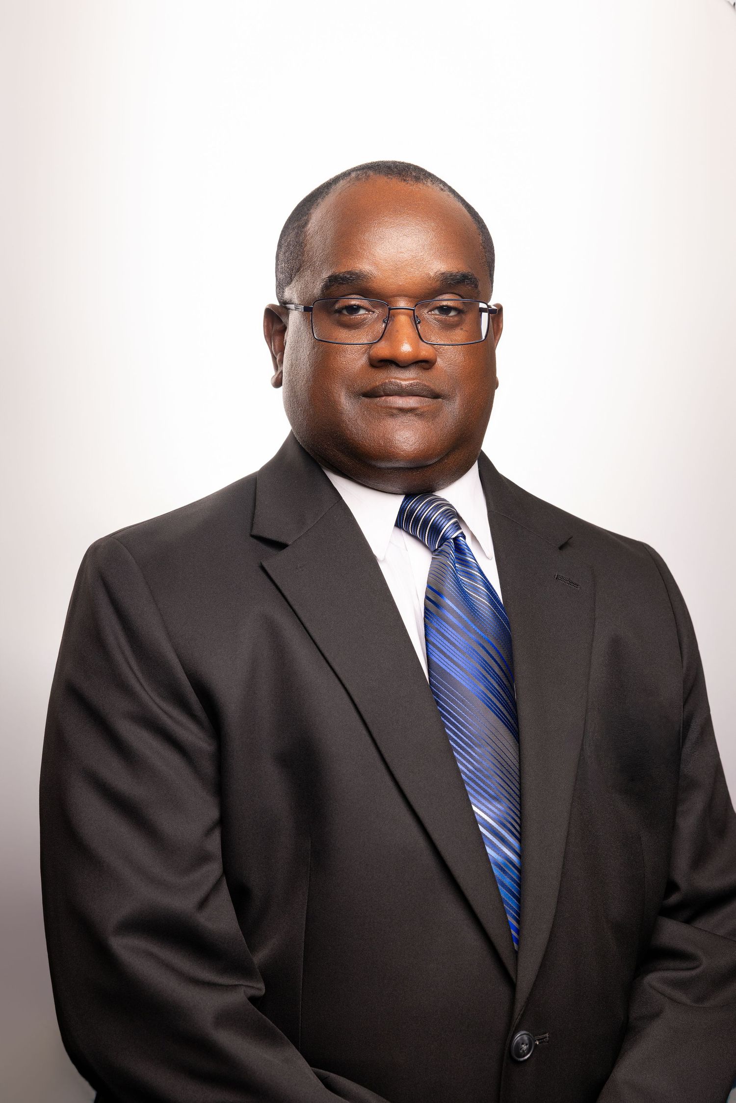 Professional man in black suit and blue striped tie wearing glasses, formal business portrait on white background.