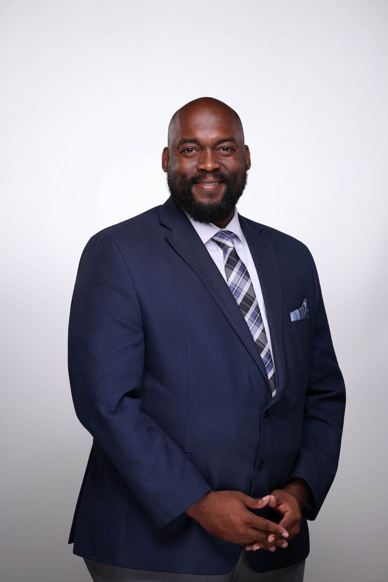 Professional man in navy suit and plaid tie smiling confidently against a light gray background.