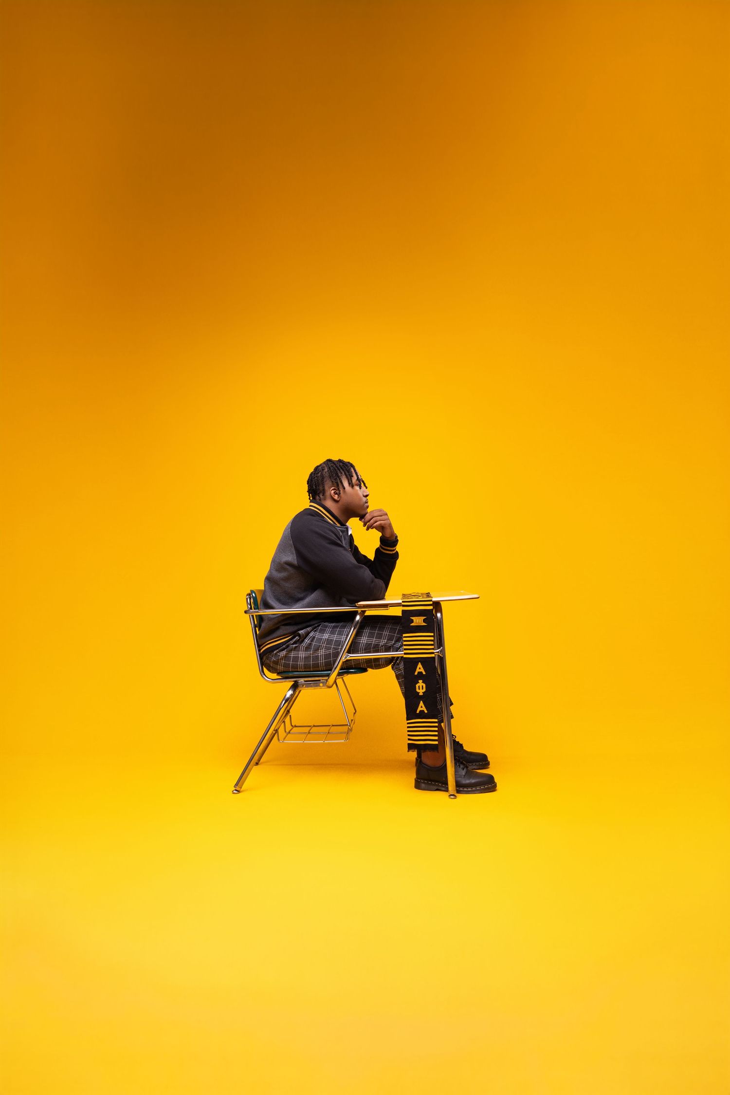 Young man sitting at a school desk in thoughtful pose against vibrant yellow background, wearing plaid pants with Greek letters.