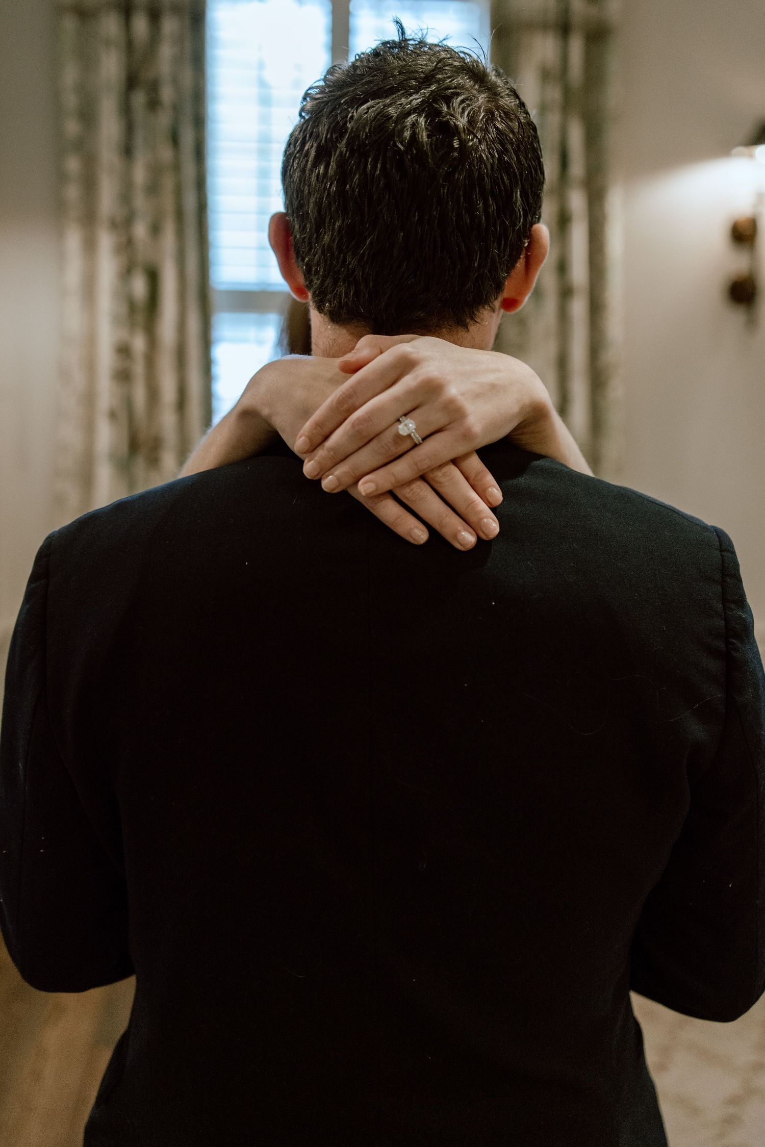 A woman with an engagement ring rests her hand on a man's neck as he stands with his back turned in a dimly lit room.