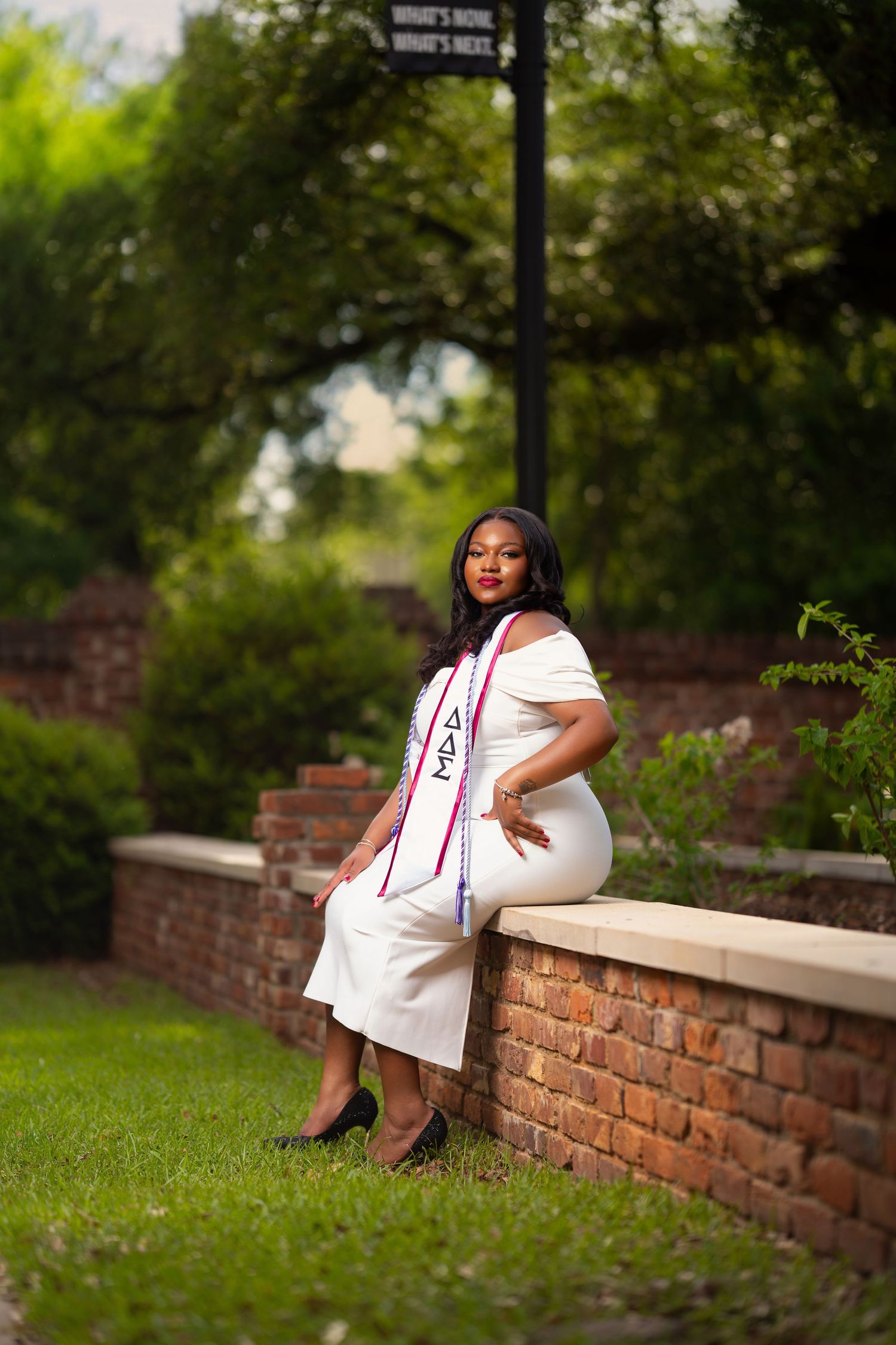 Young woman in white dress and graduation sash poses on brick wall outdoors for senior portrait.