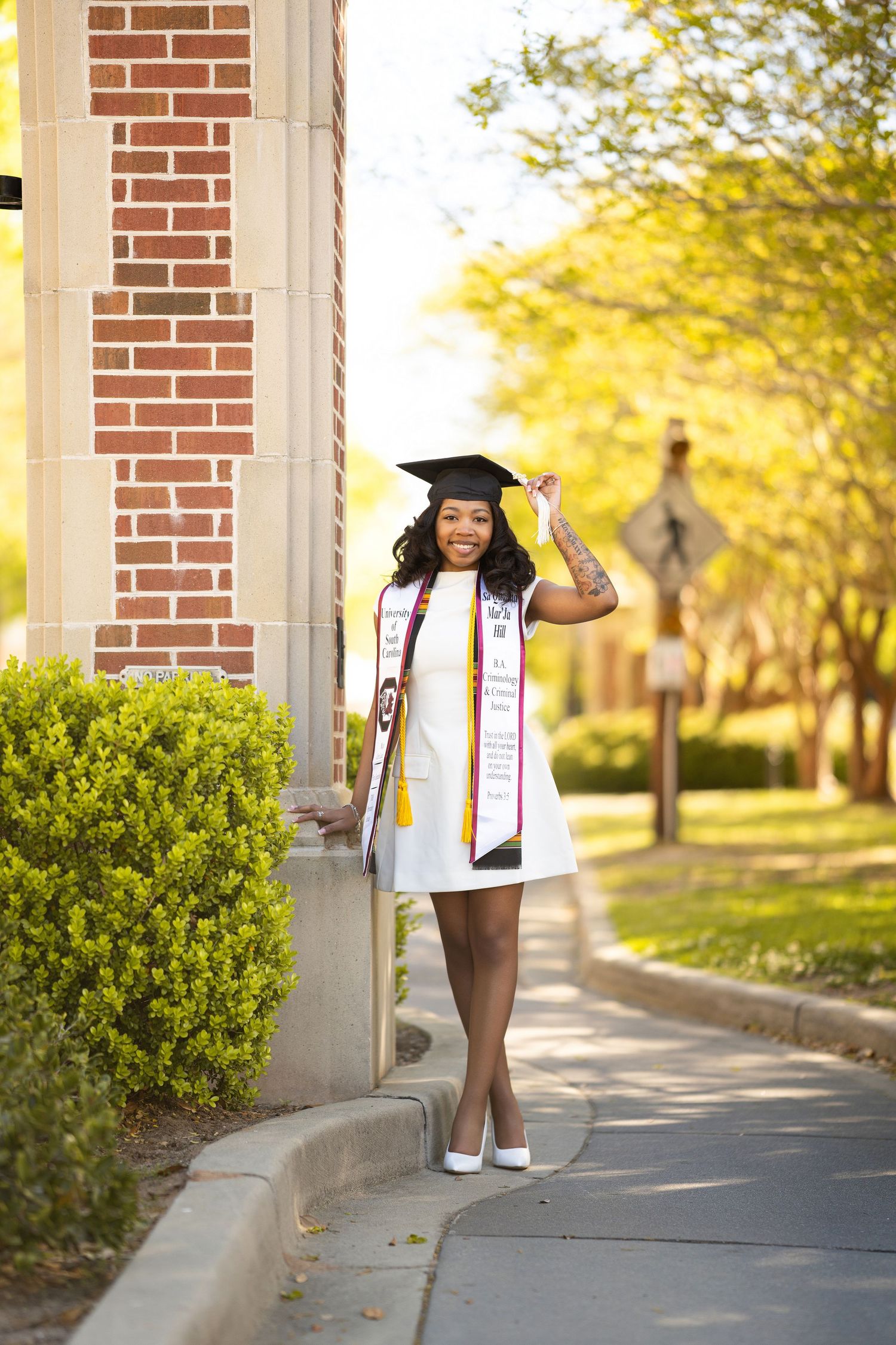 Young woman in white graduation gown and cap poses proudly on a sunny campus walkway with brick buildings.