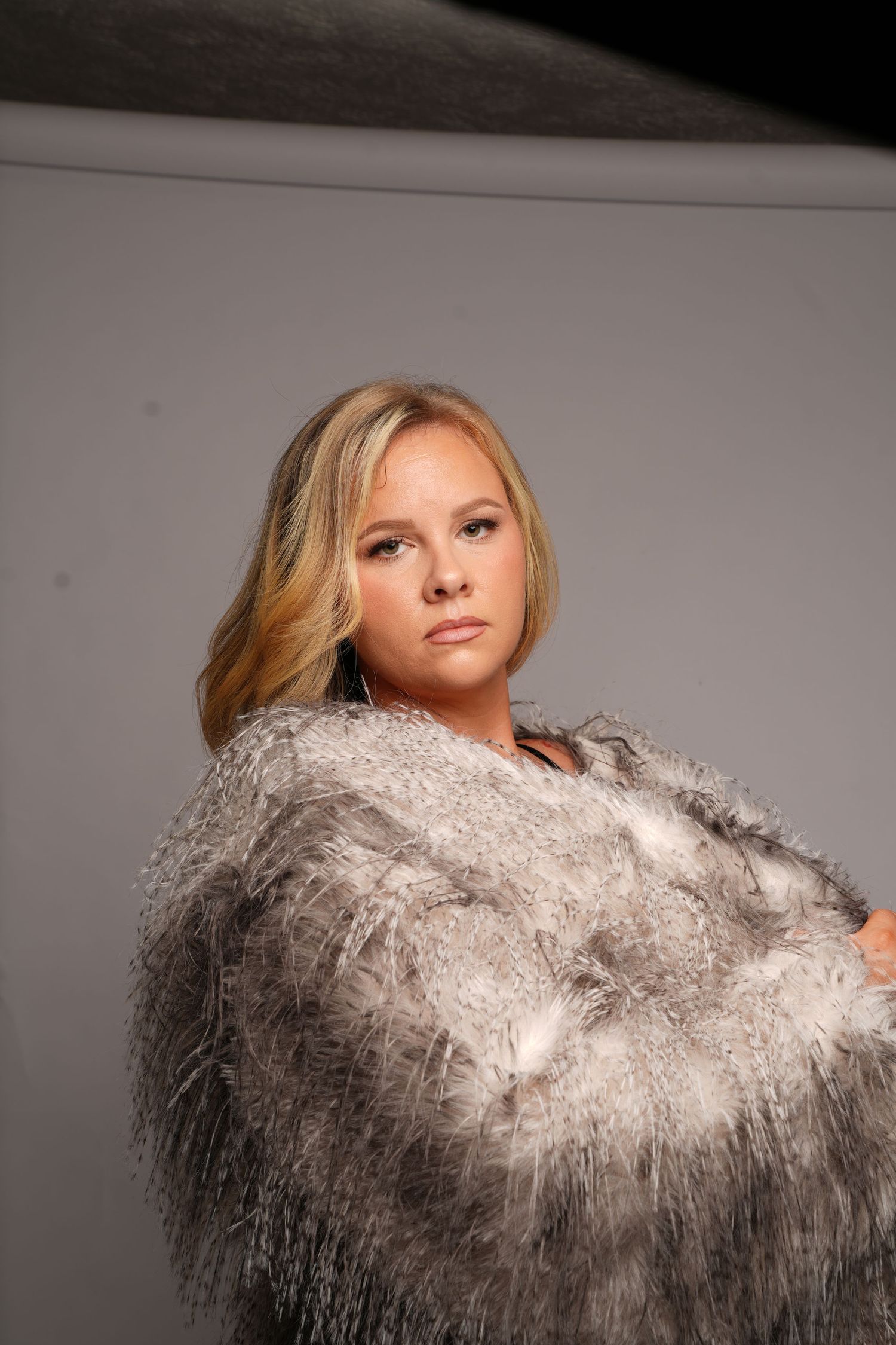 Young blonde woman posing in a fluffy gray fur coat against a neutral studio background.