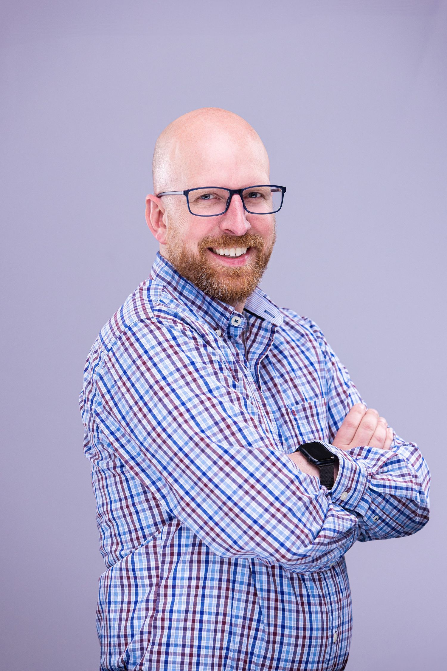 Professional headshot of person in checkered shirt with crossed arms and glasses against purple backdrop.