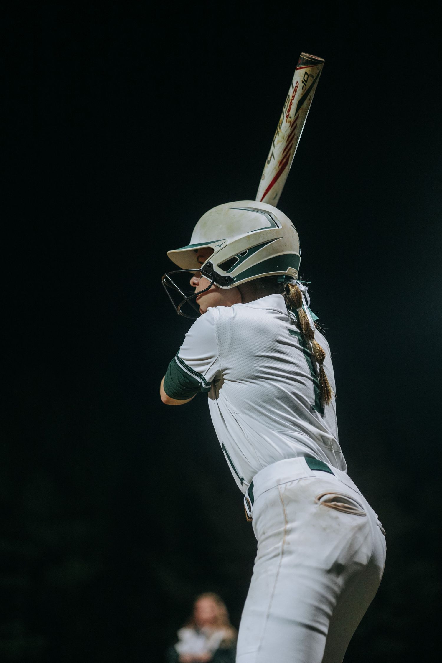 Baseball batter in white uniform stands at the plate against dark background during night game.