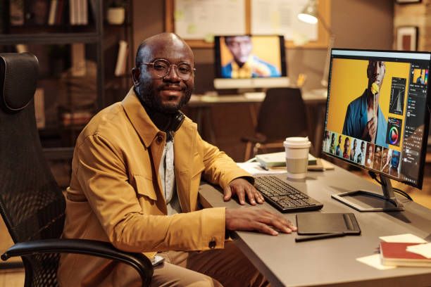Professional in a yellow blazer works at a computer desk setup with multiple monitors in a modern office environment.