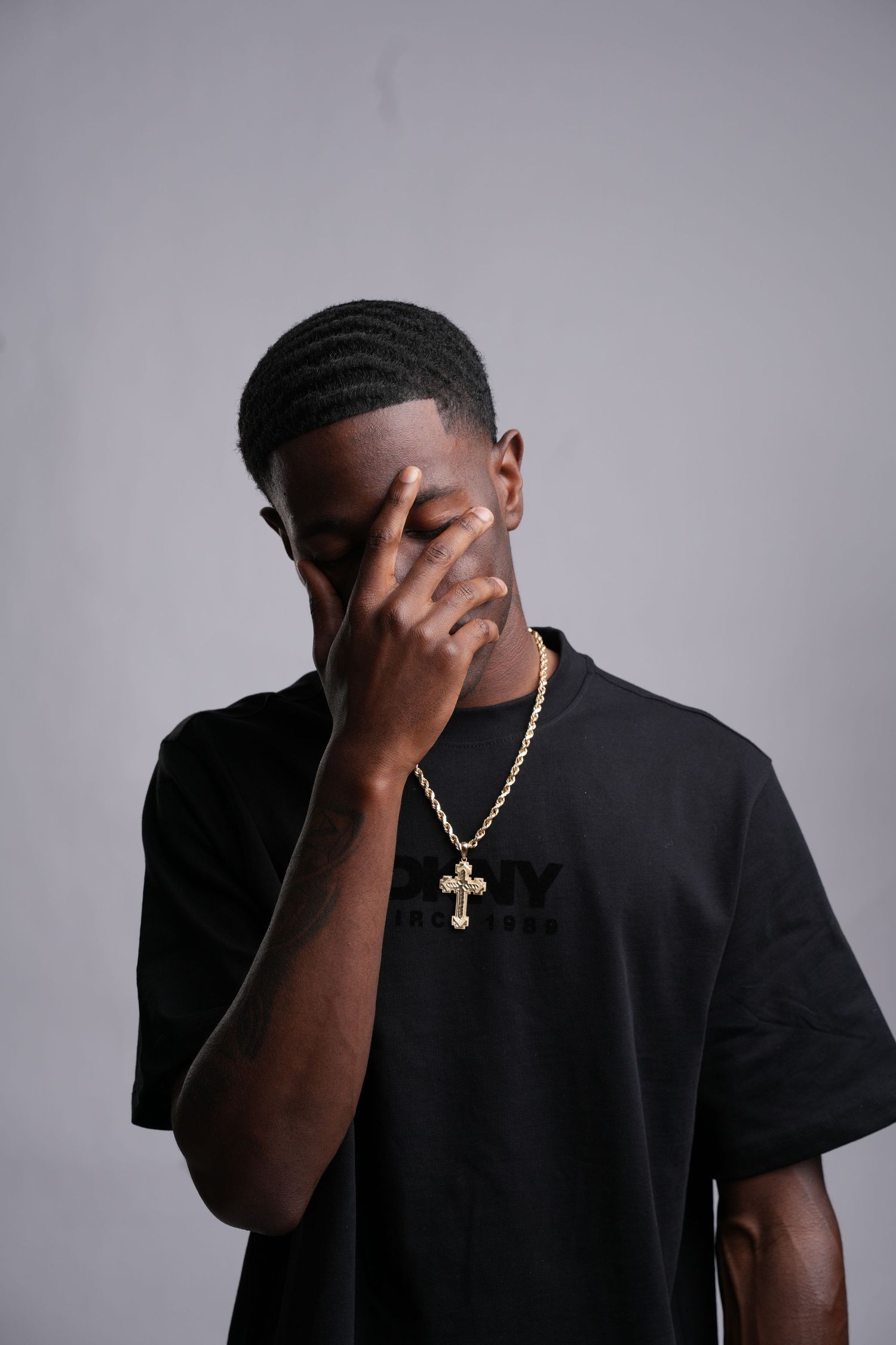 Young man in black oversized tee with gold cross necklace, hand covering face, posing against gray background.