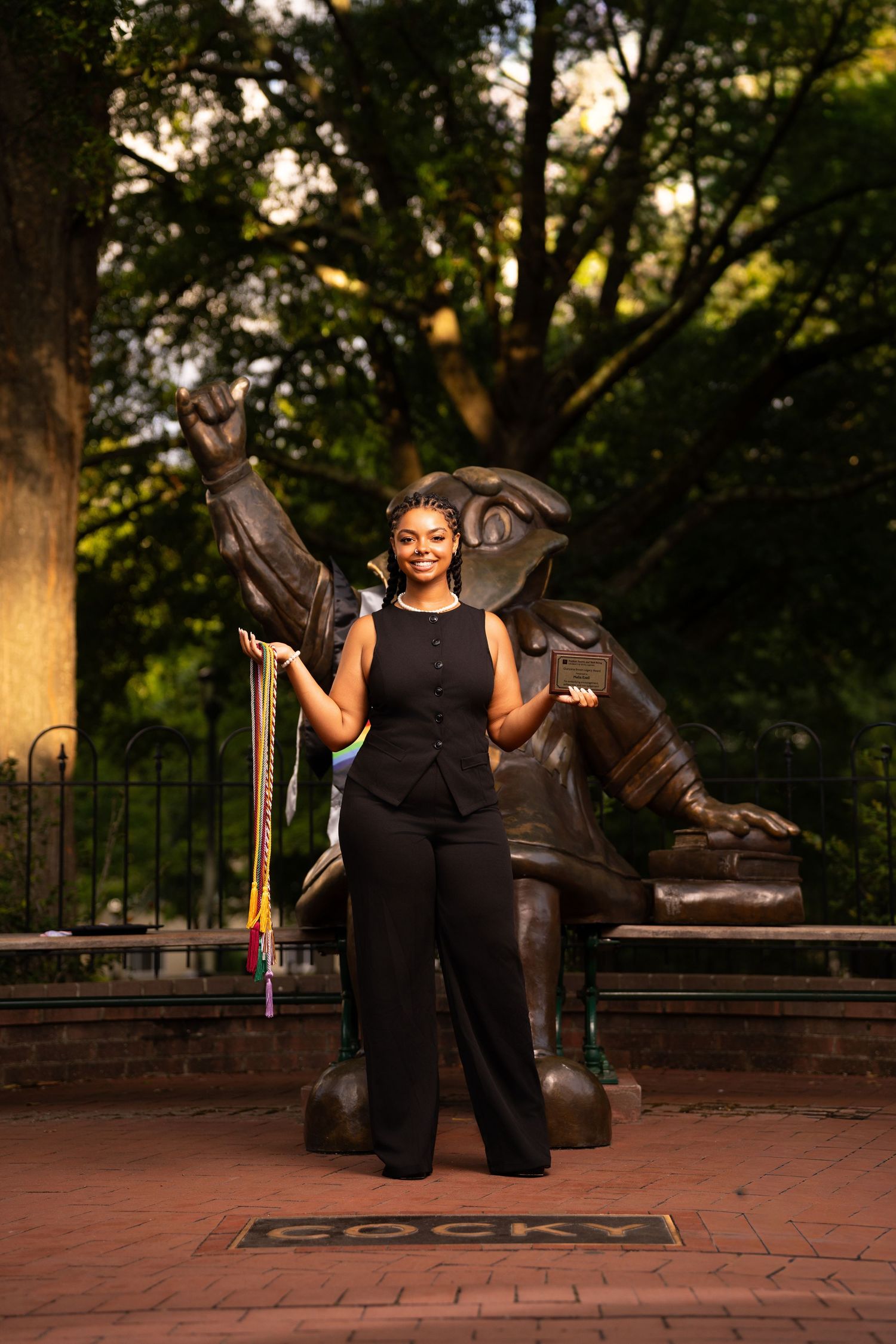 A graduate poses proudly in black attire holding colorful cords before the Cocky statue outdoors.
