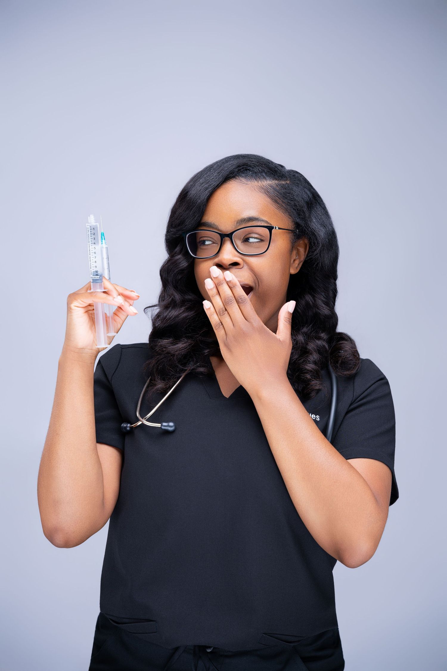A Black female nurse in black scrubs and glasses holds a syringe with a surprised expression, hand over mouth.