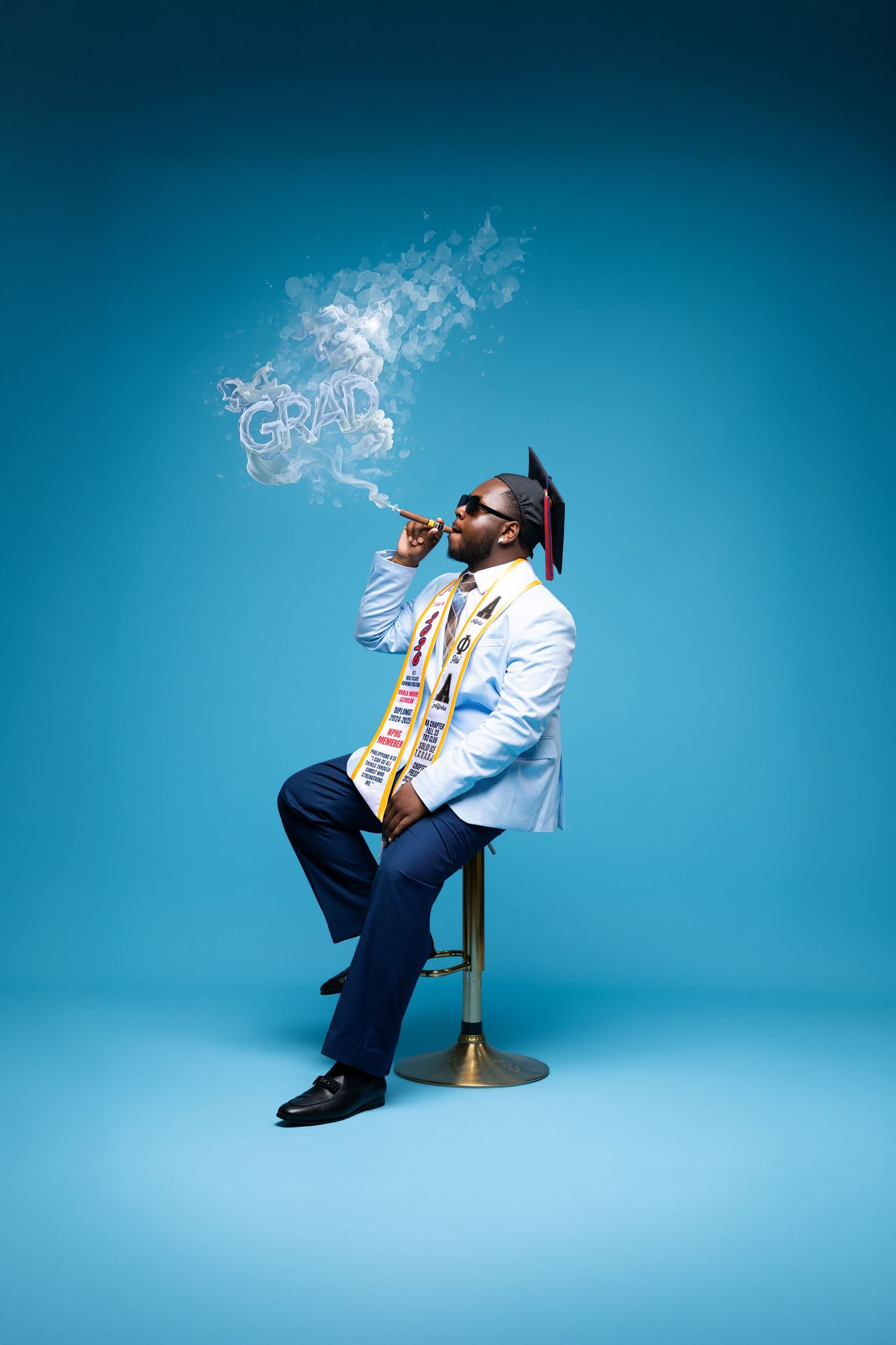 Graduate in white blazer and cap blowing smoke rings spelling GRAD on blue background.