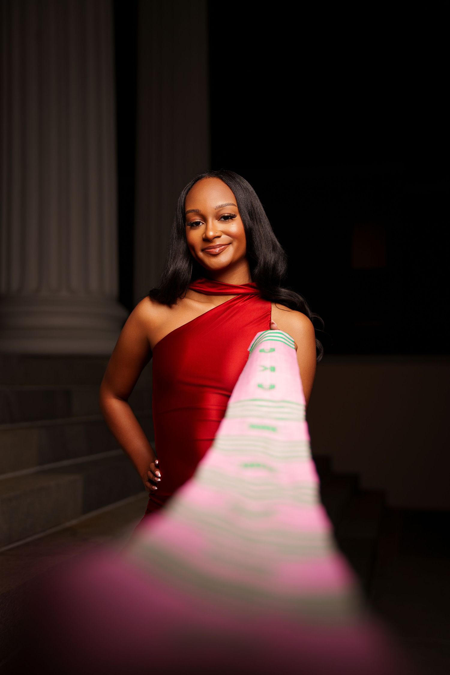 Woman in red one-shoulder gown smiling elegantly on steps with dramatic lighting and architectural columns.