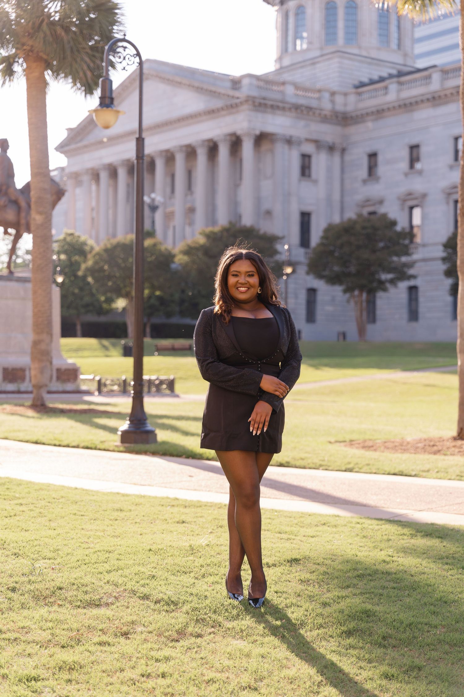 Young woman in black dress smiling outdoors near a government building with columns and palm trees.