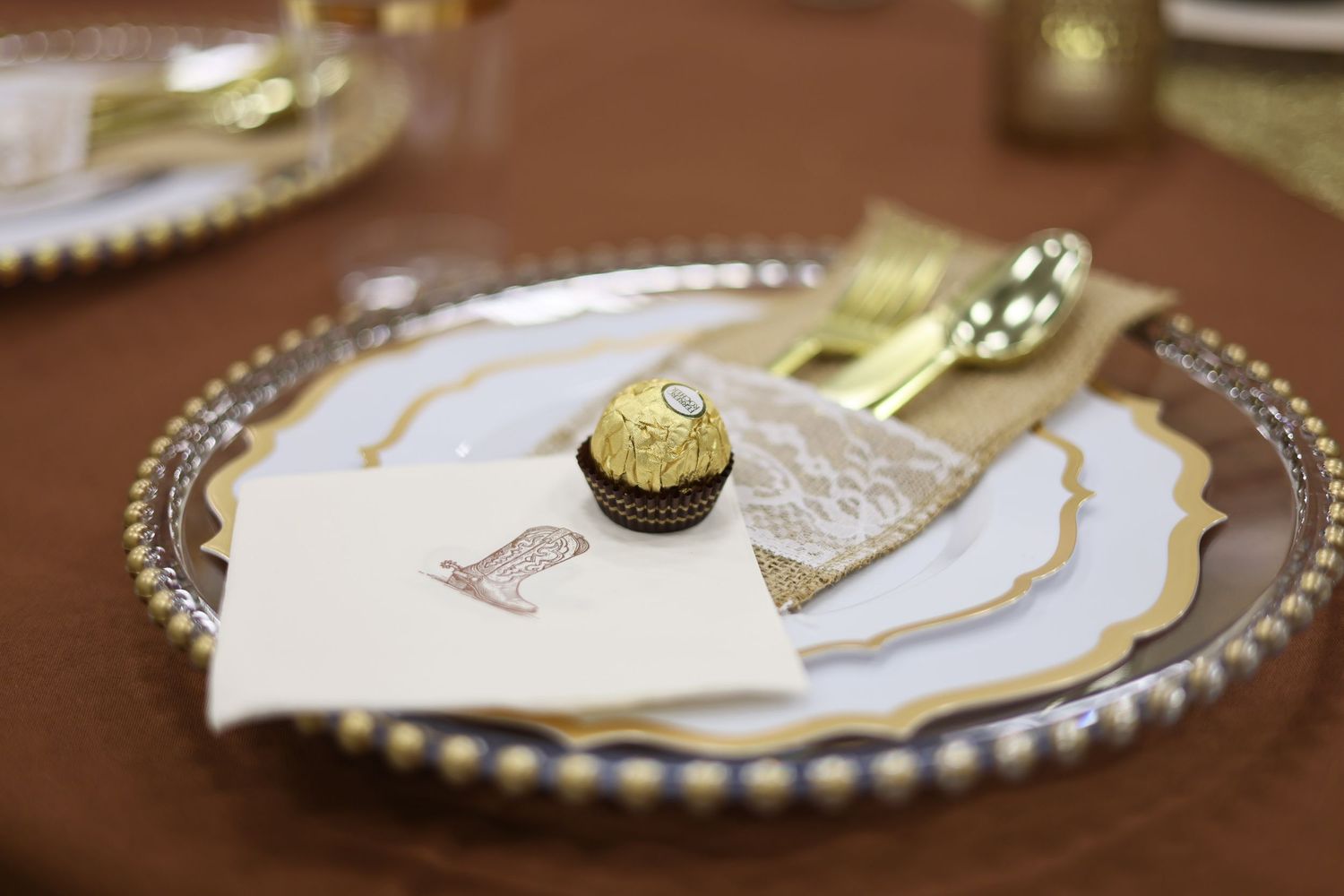 Elegant place setting with a Ferrero Rocher chocolate on white and gold plates with golden cutlery on a brown tablecloth.