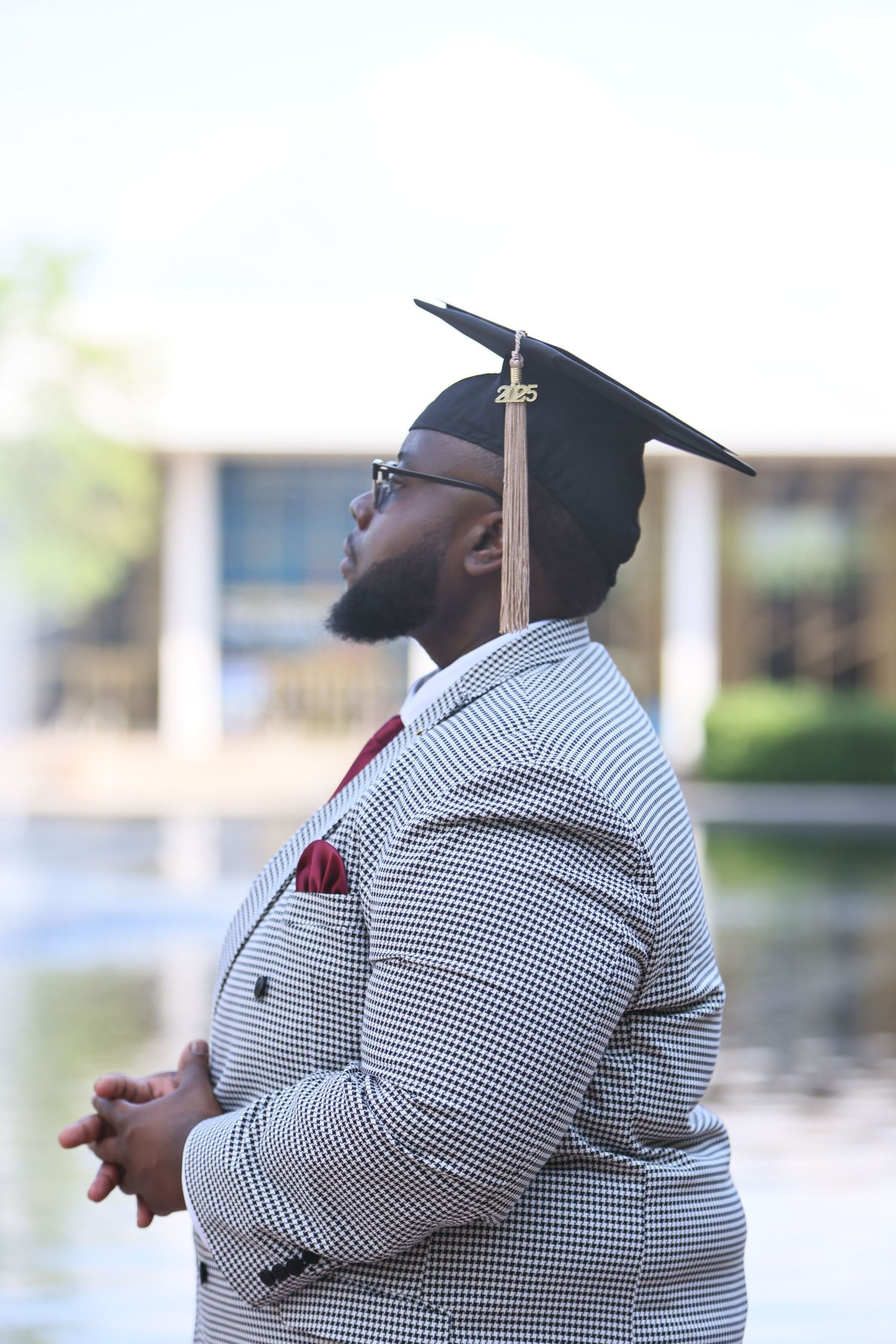 Graduate in cap and gown reflecting near a tranquil pond.