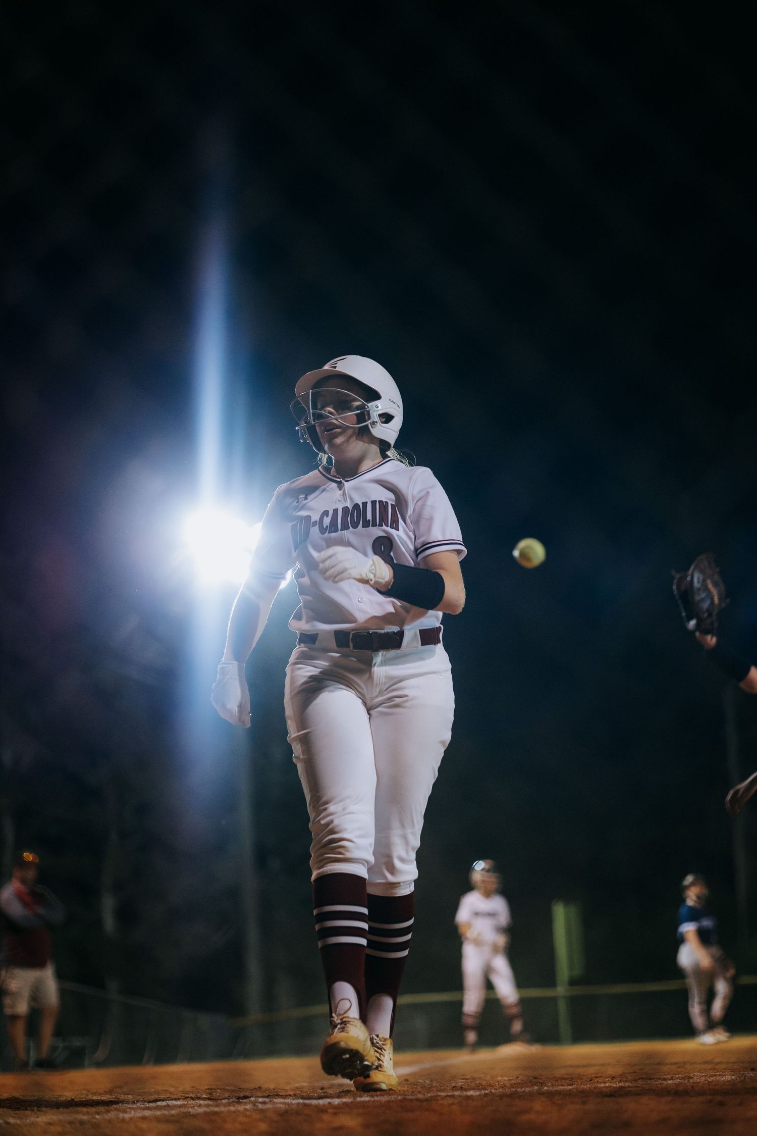 Softball pitcher in white uniform mid-delivery under stadium lights at night.
