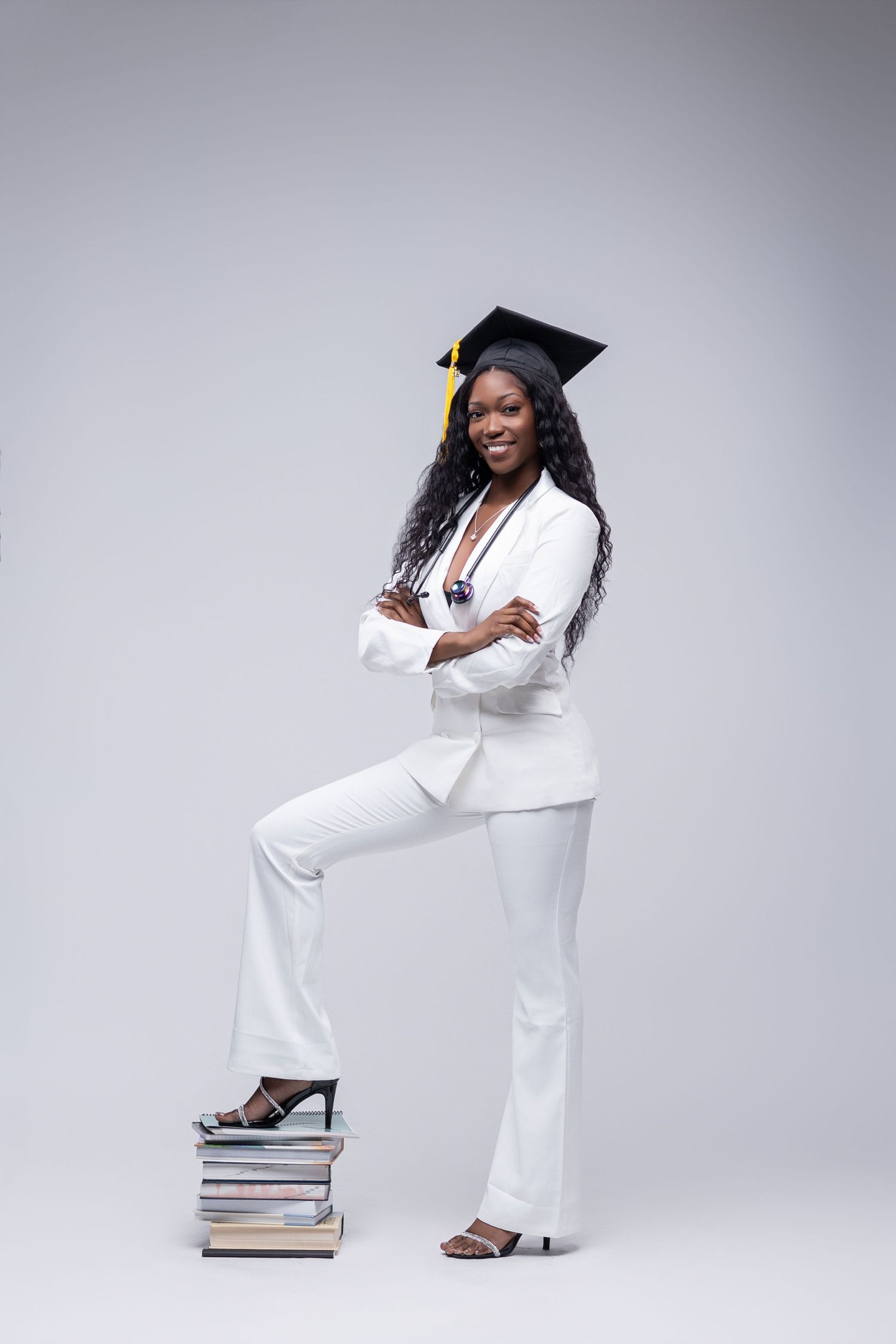 Graduate in white suit stands confidently with foot on stack of books against gray background.