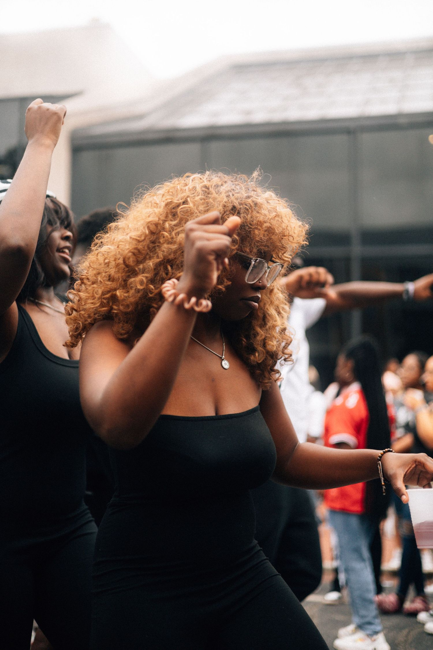 A dancer in a black top with curly hair performs an expressive dance movement with arms raised.