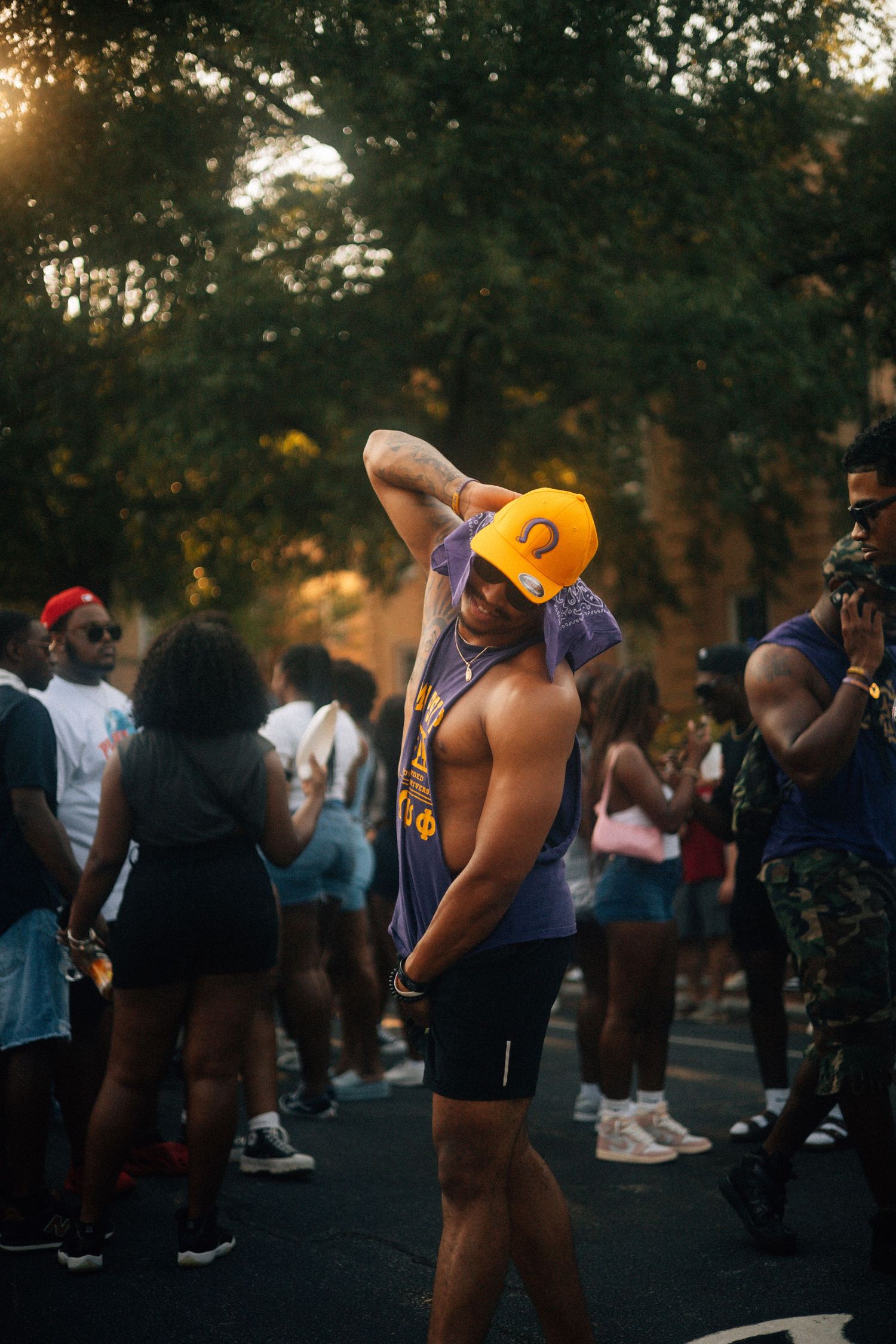 Person in yellow cap and purple tank top poses during an outdoor social gathering at sunset.