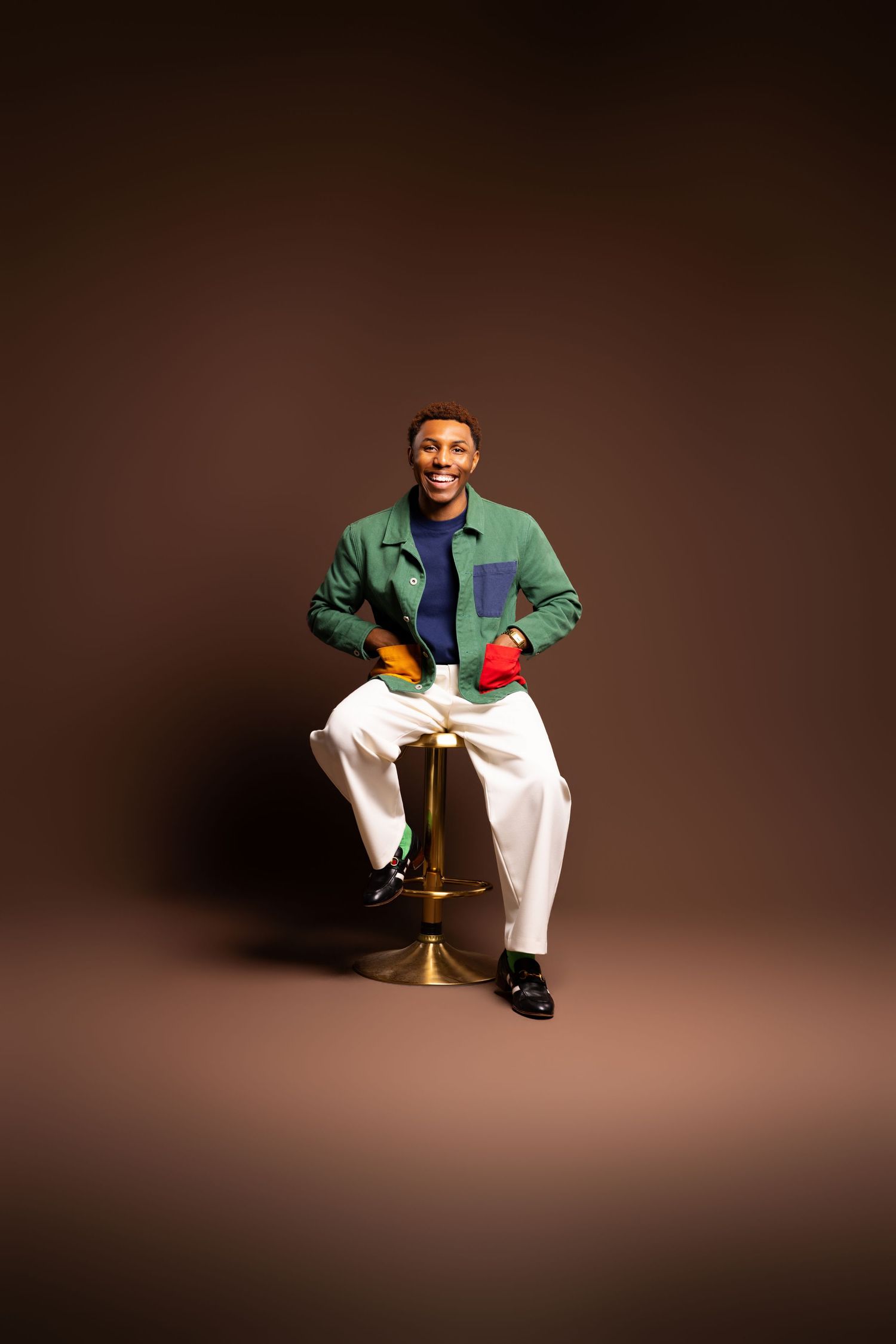 Person in a green jacket and white pants sits casually on a bar stool against a dark brown studio backdrop.