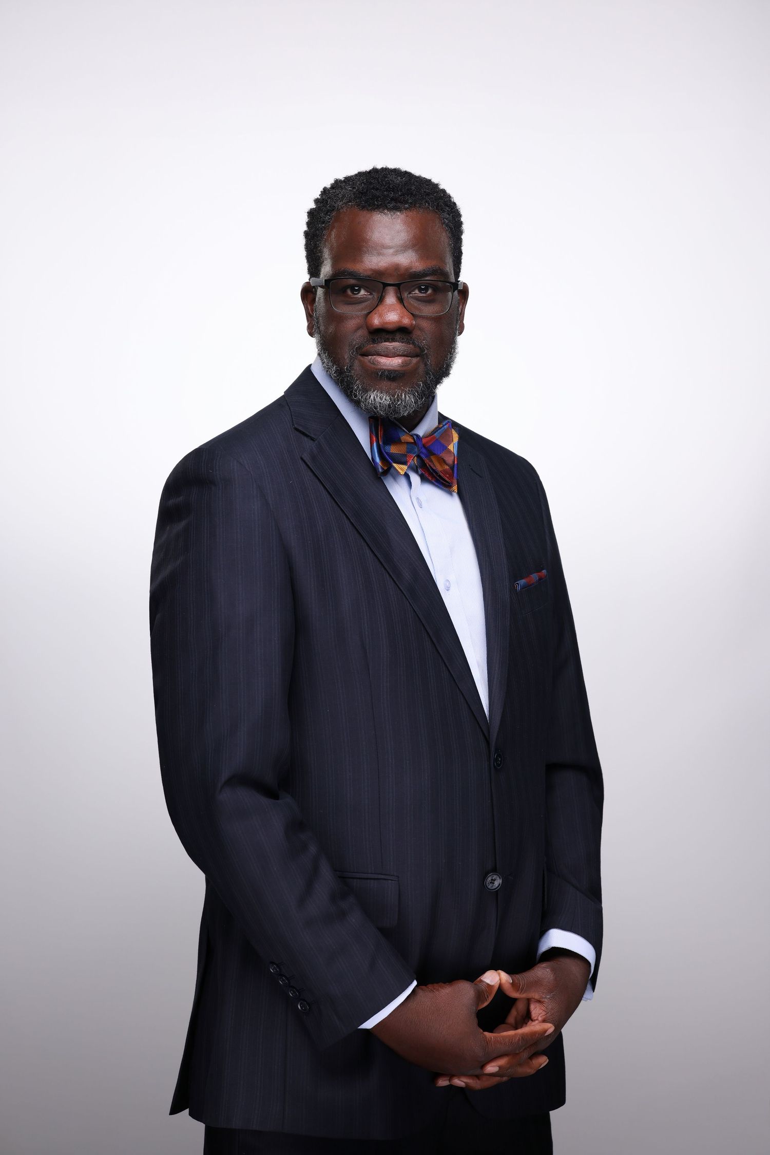 Professional man in navy pinstripe suit and colorful bow tie with glasses poses for formal portrait.