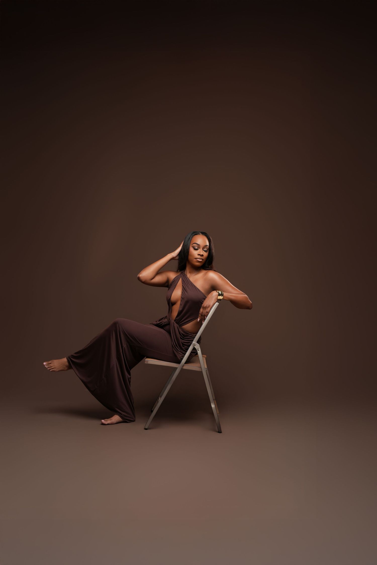A Black woman in a brown halter jumpsuit poses gracefully on a folding chair against a dark brown studio background.