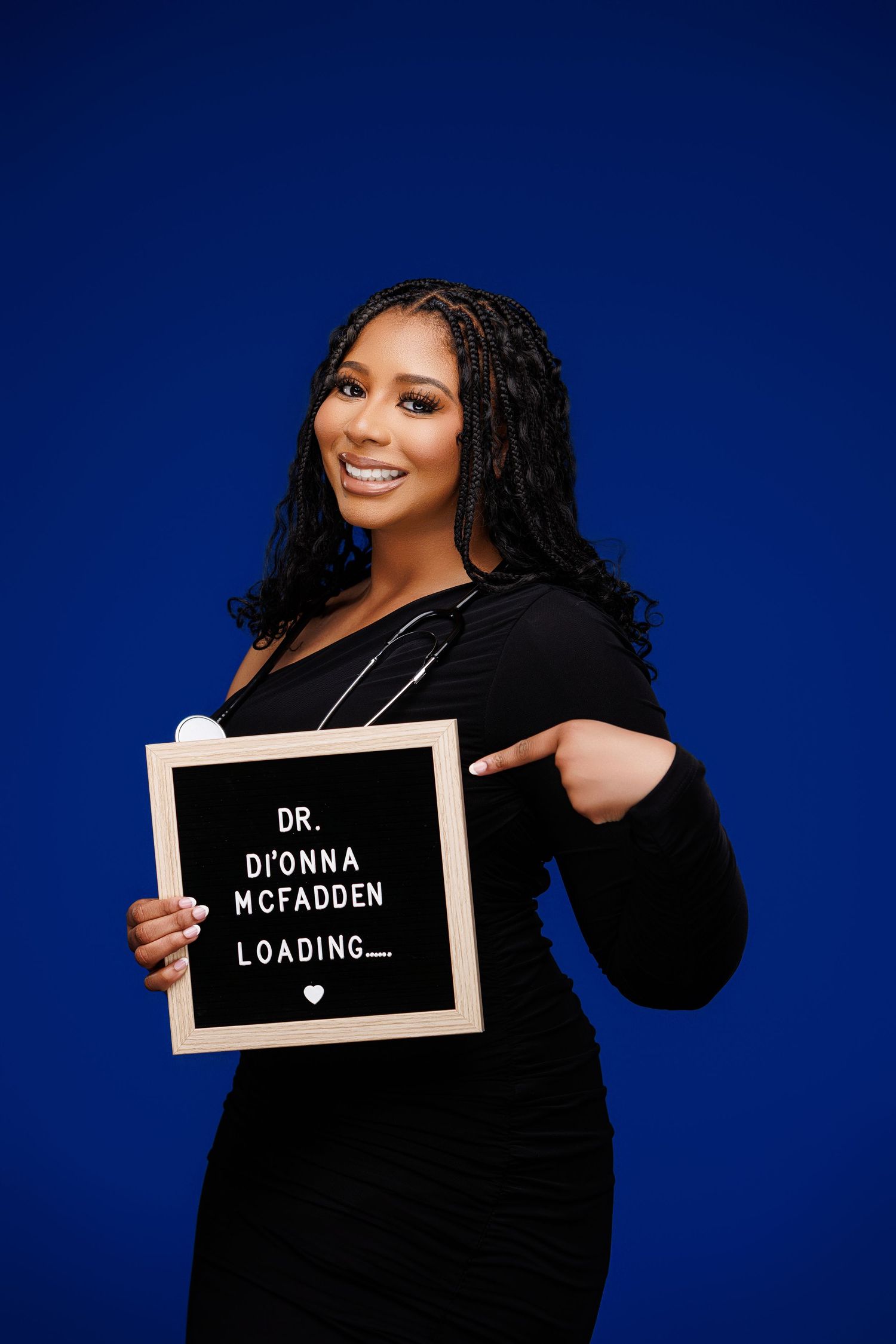 Woman in black dress holding a letter board sign reading Dr. DiOnna McFadden Loading, wearing a stethoscope.