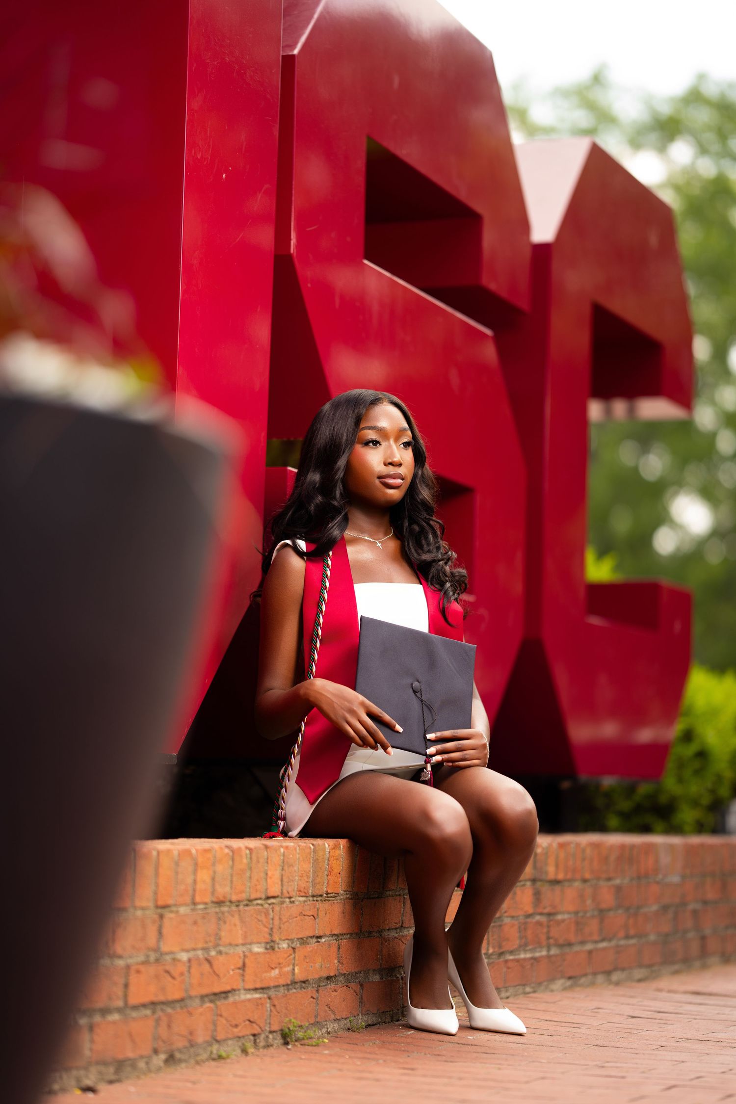 Young Black woman in graduation attire sits before red university sculpture, holding cap and diploma.